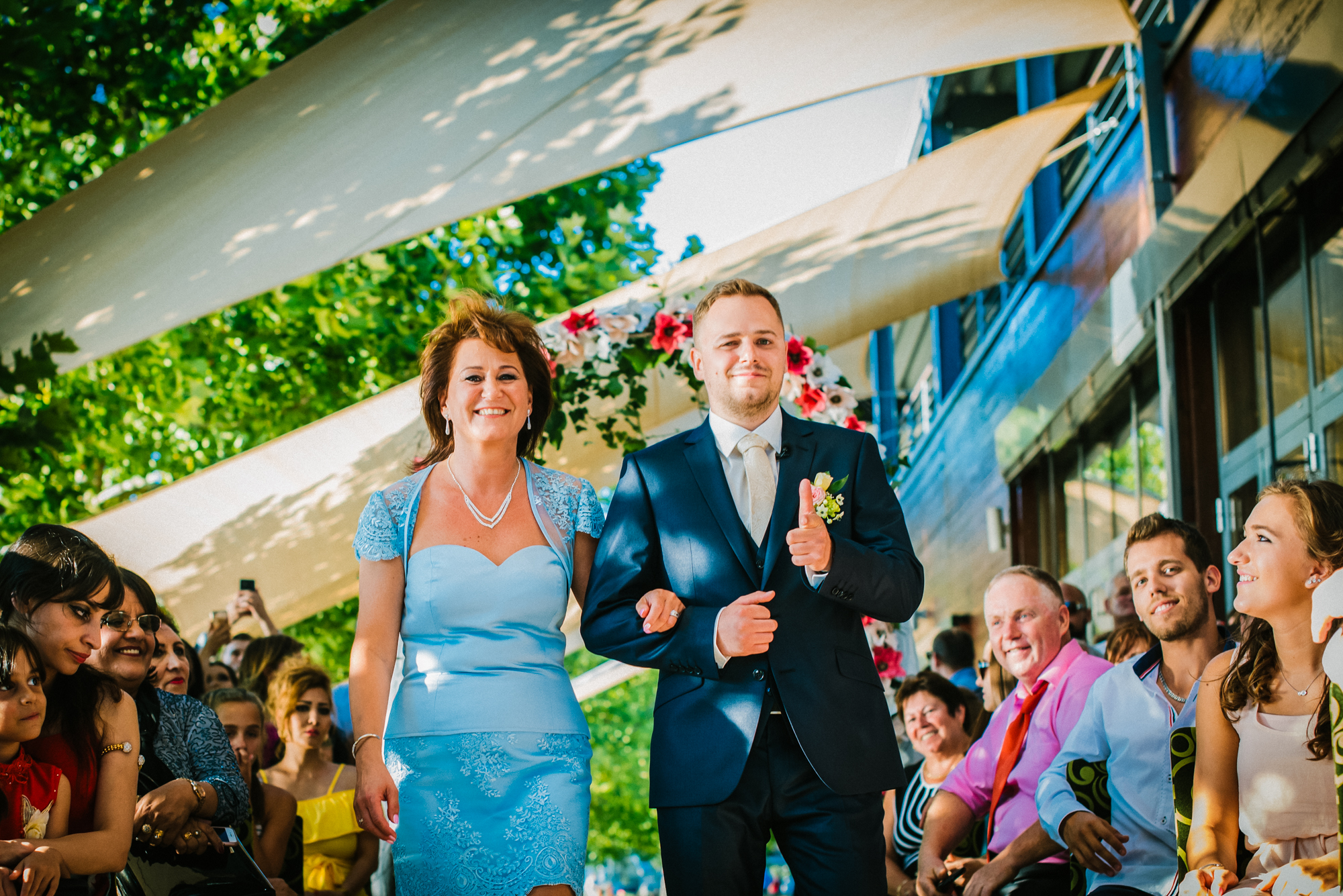Groom walking with his mum down the aisle, in bright natural light.