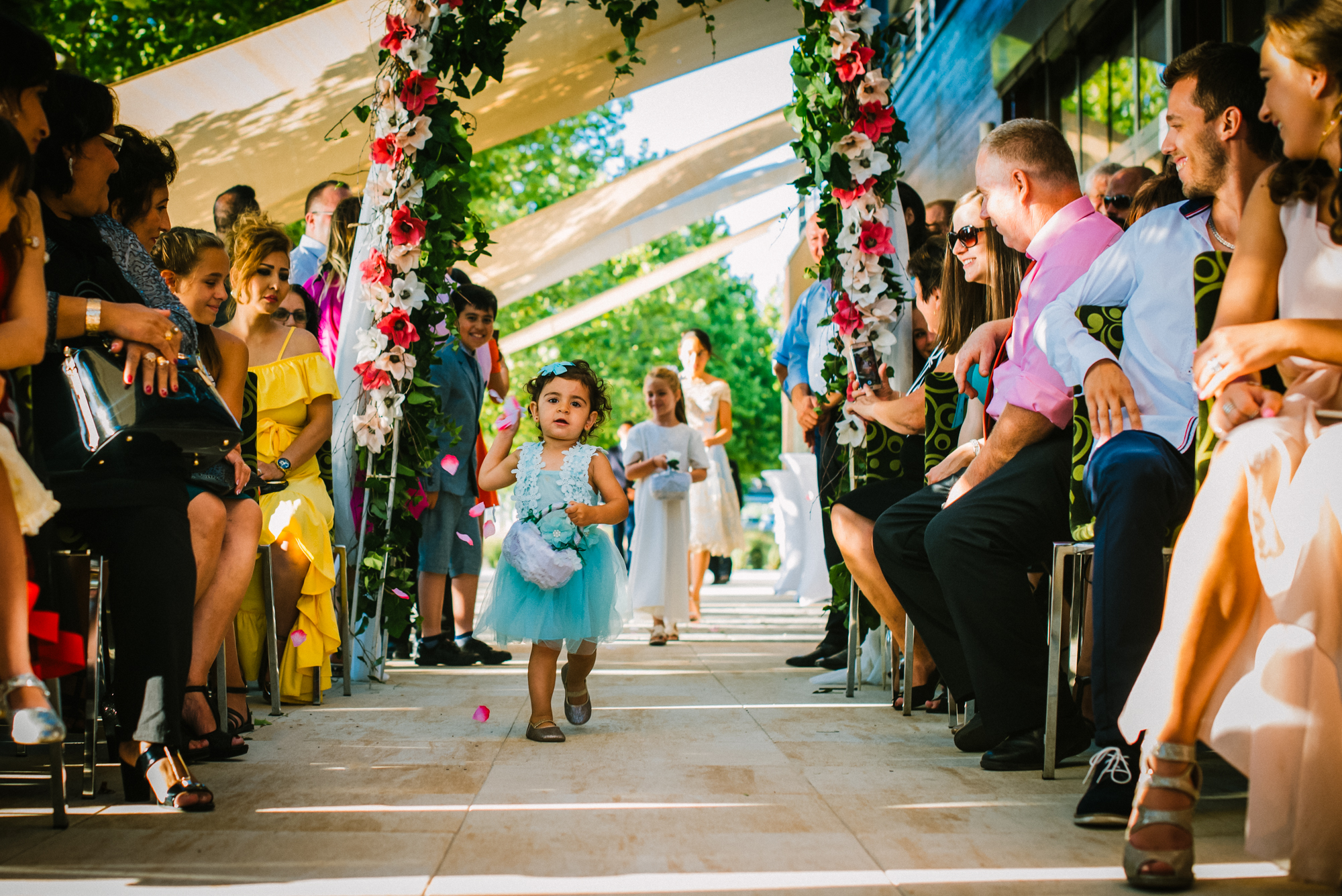 Toddler flower girl throwing petals during outdoor multicultural wedding ceremony in Budapest.