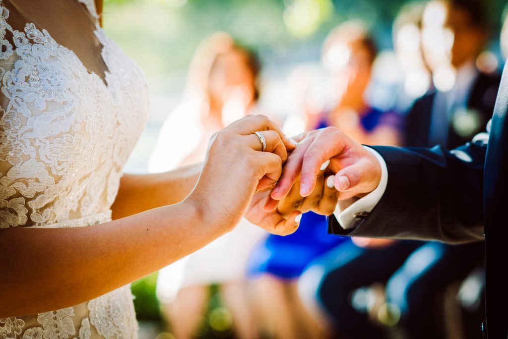 Close-up of wedding ring exchange during outdoor ceremony with blurred guests in background.