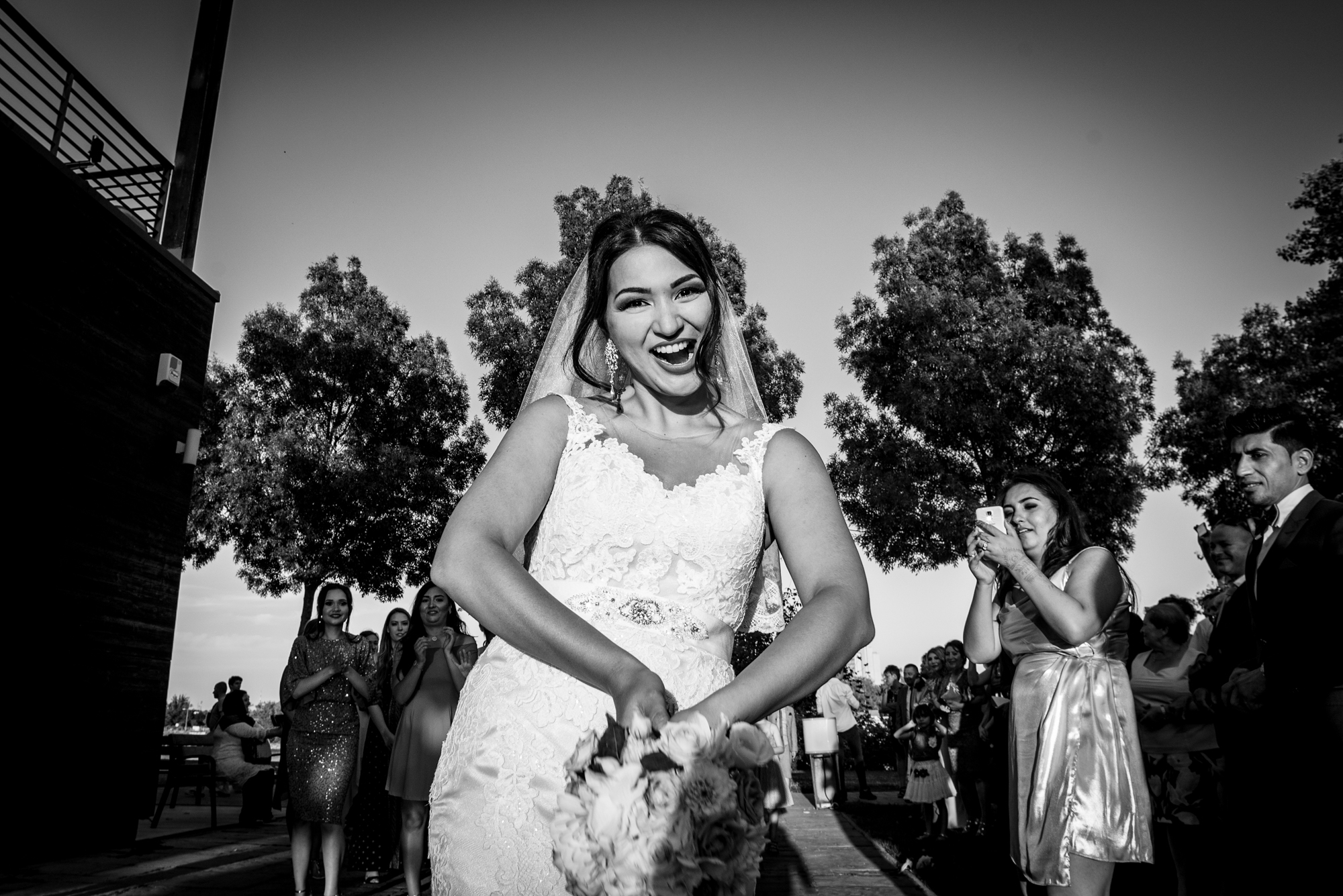 Bride smiling before bouquet toss with guests gathered outdoors at Budapest wedding celebration.