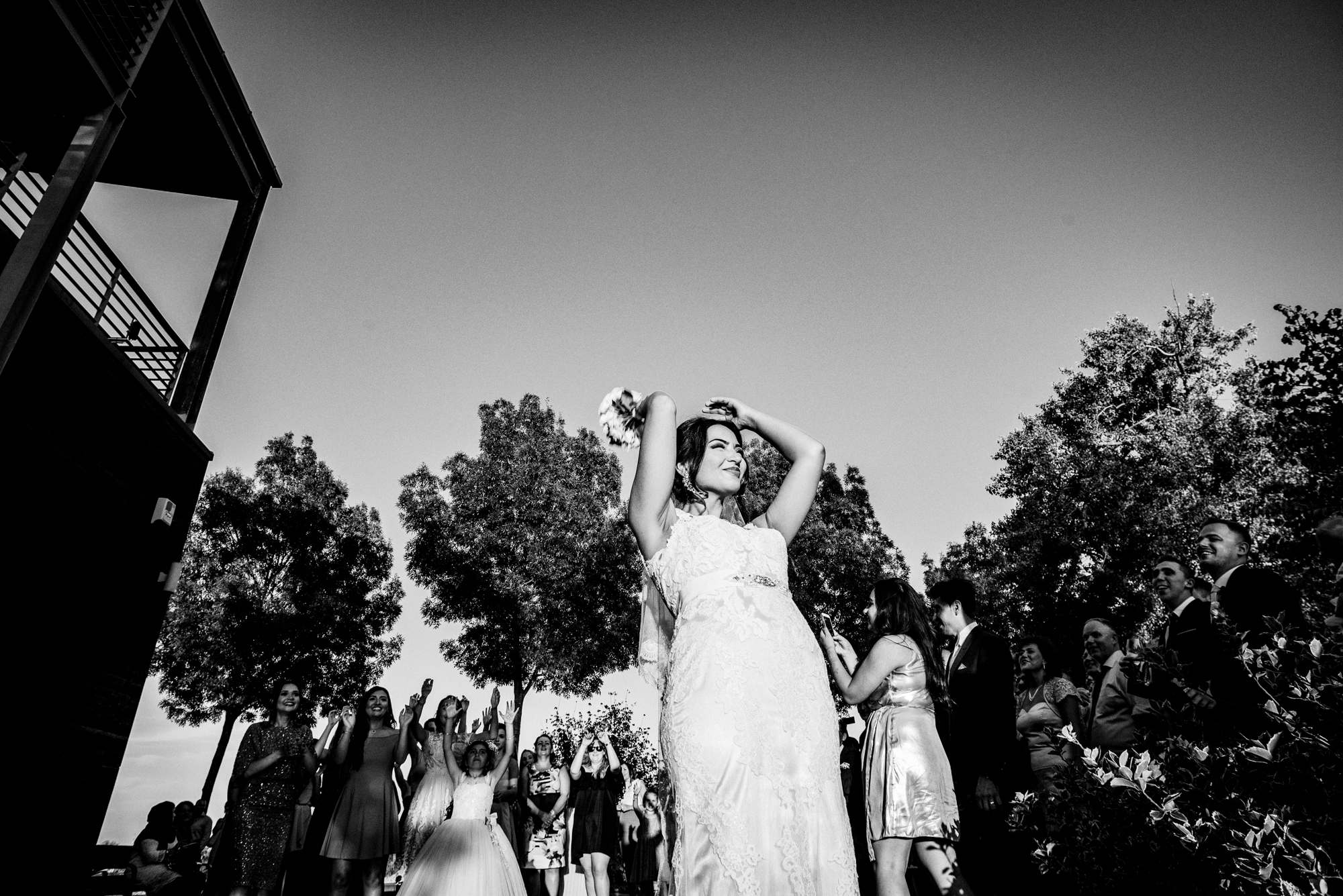 Bride lifting bouquet above her head surrounded by cheering wedding guests during outdoor reception.