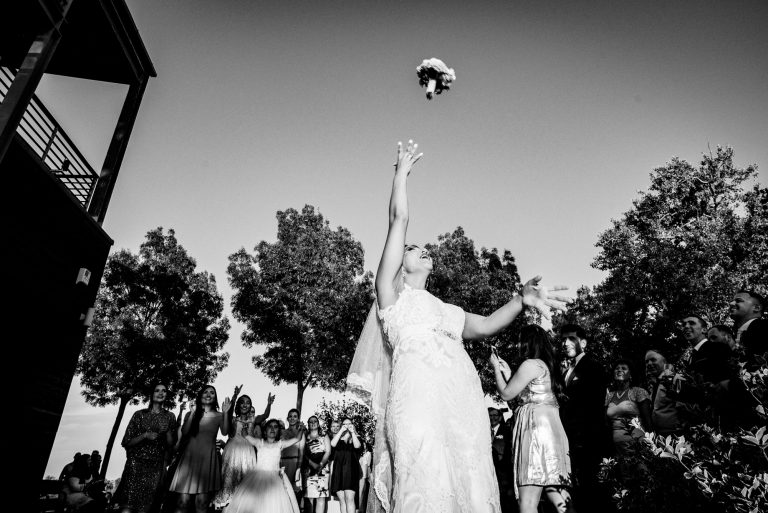 Bouquet flying through the air as wedding guests reach up during lively outdoor celebration.