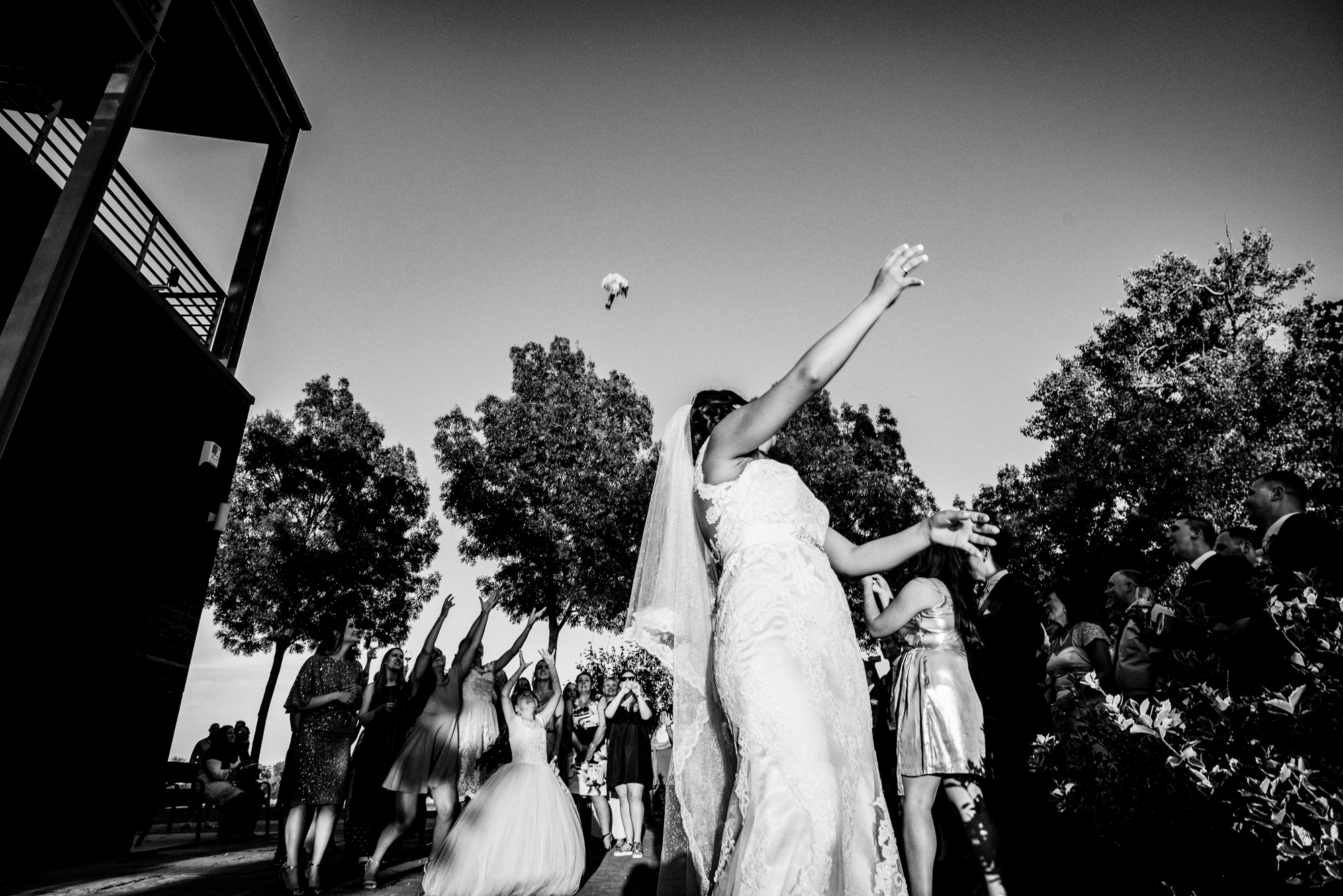 Bride turning after bouquet toss while guests react with raised hands and laughter.