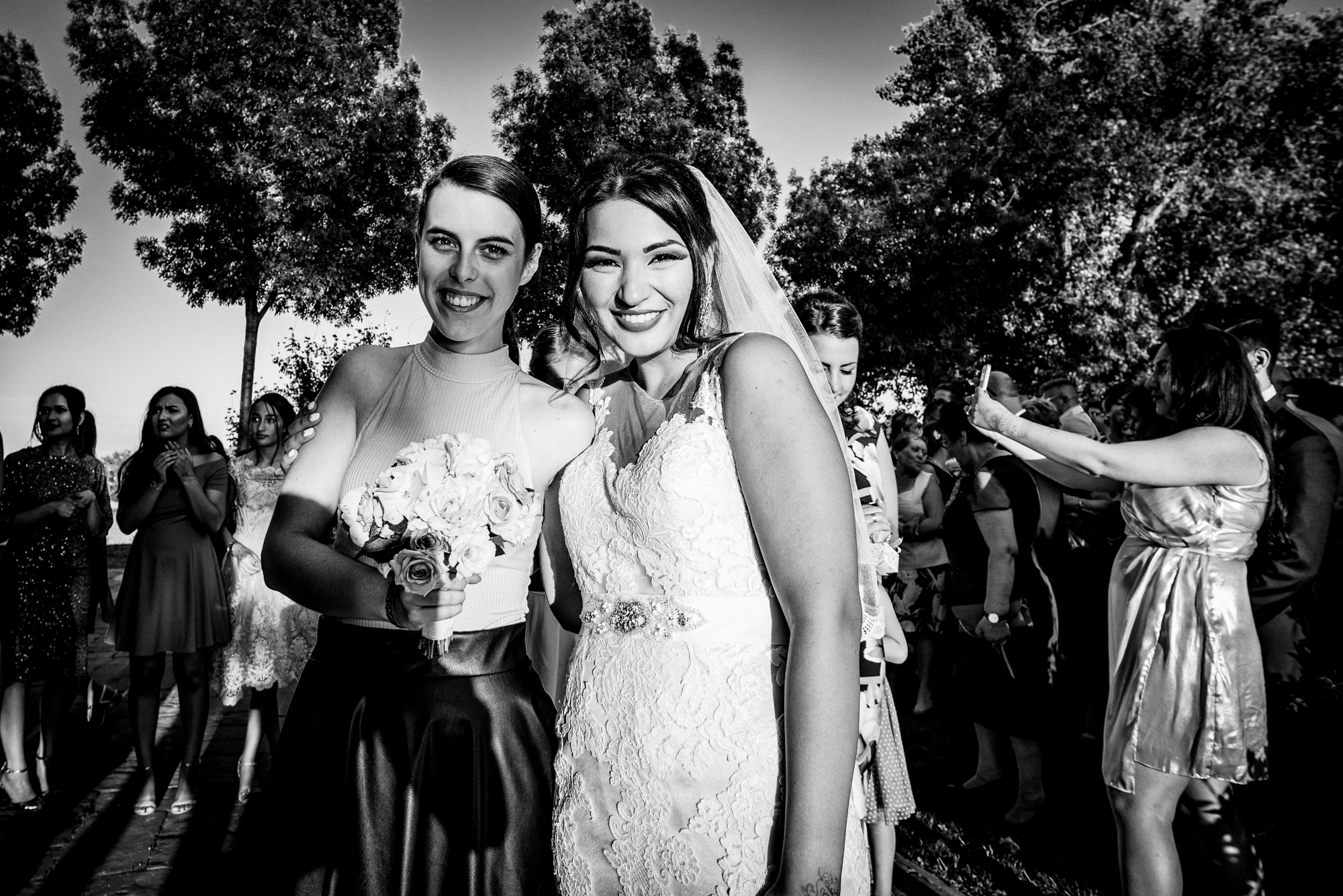 Bride and bridesmaid smiling together holding bouquet during candid wedding moment outdoors.