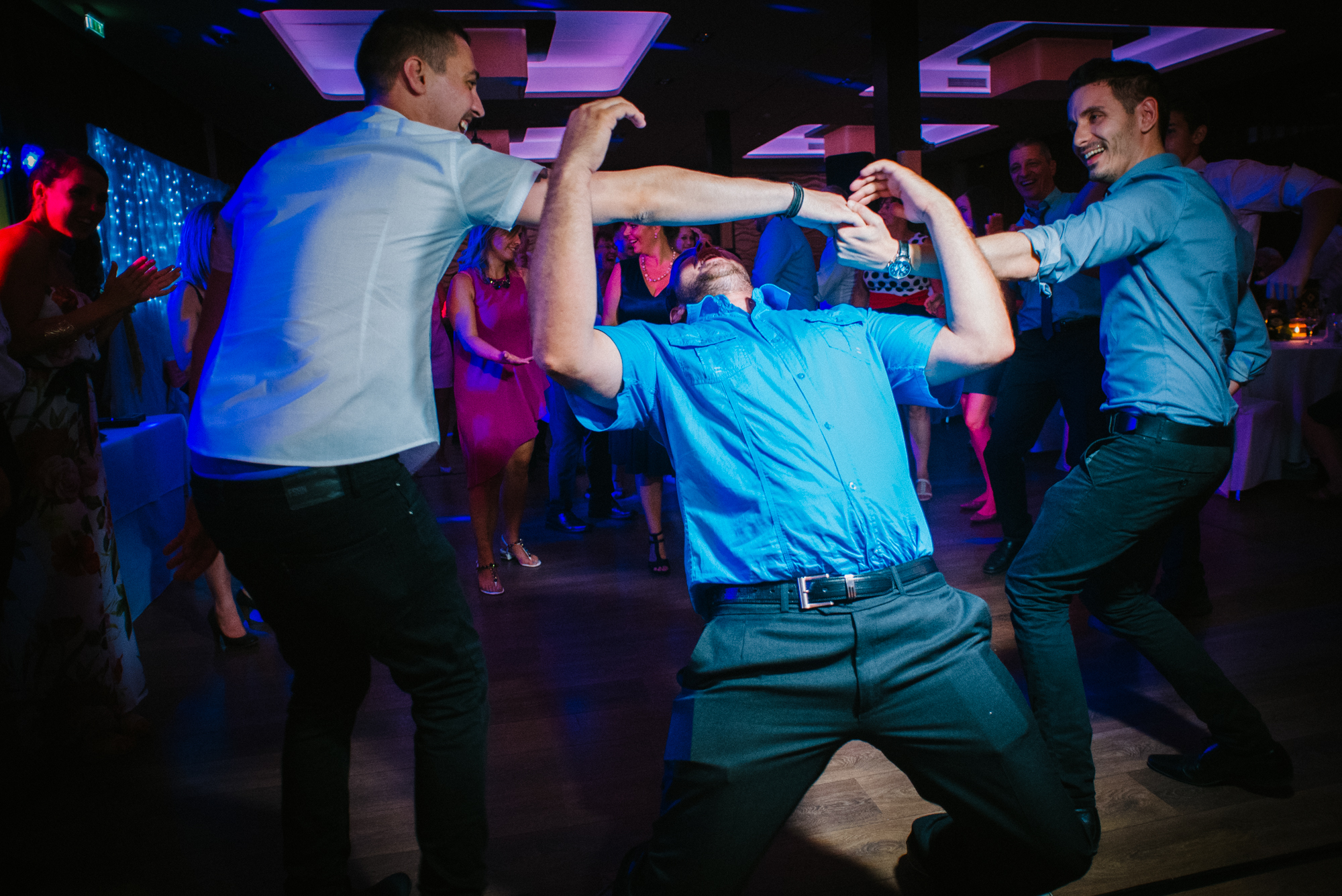 Groomsmen dancing limbo energetically in circle with dramatic lighting during wedding party in Budapest.