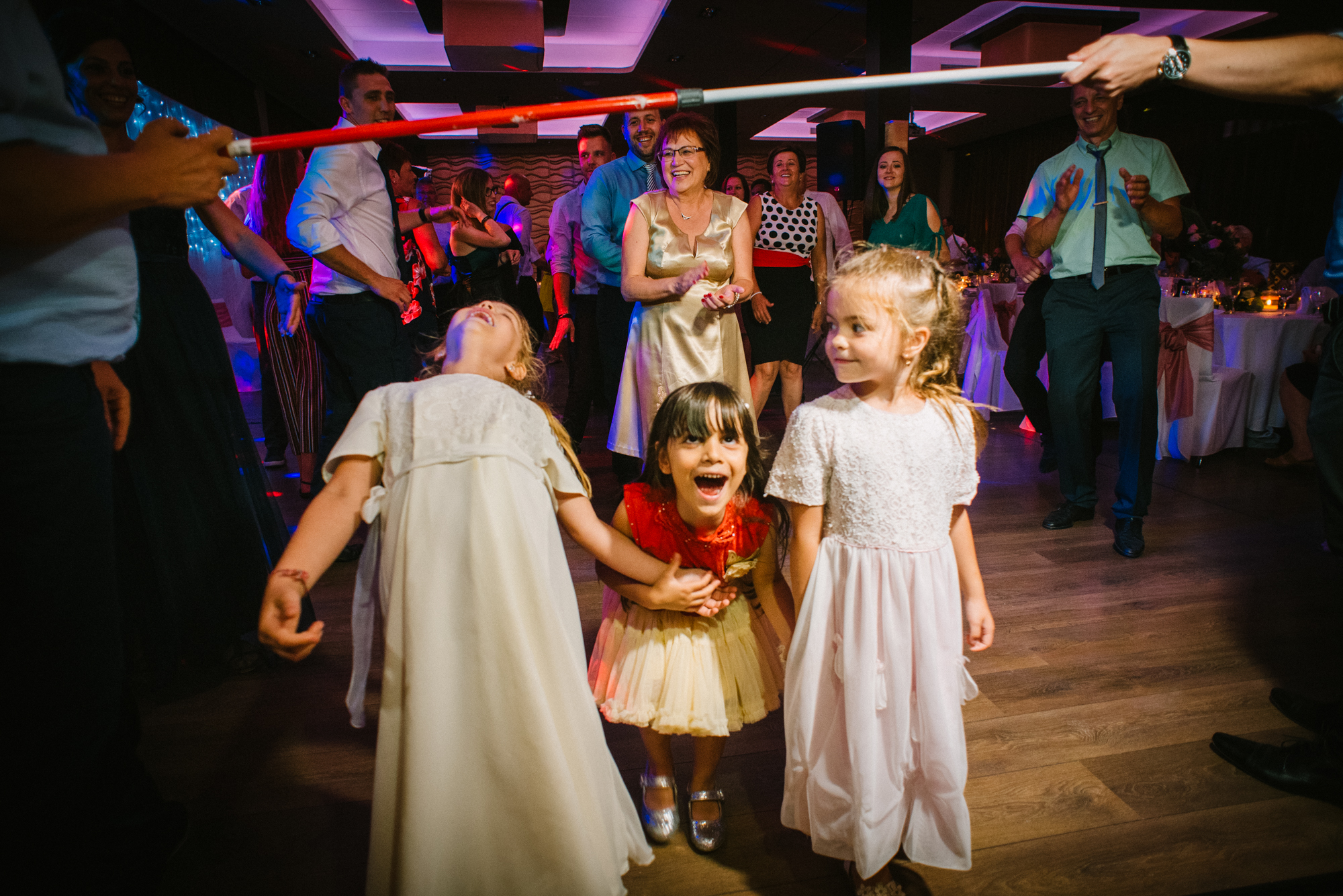 Children dancing limbo and laughing together on wedding dance floor with guests watching behind.