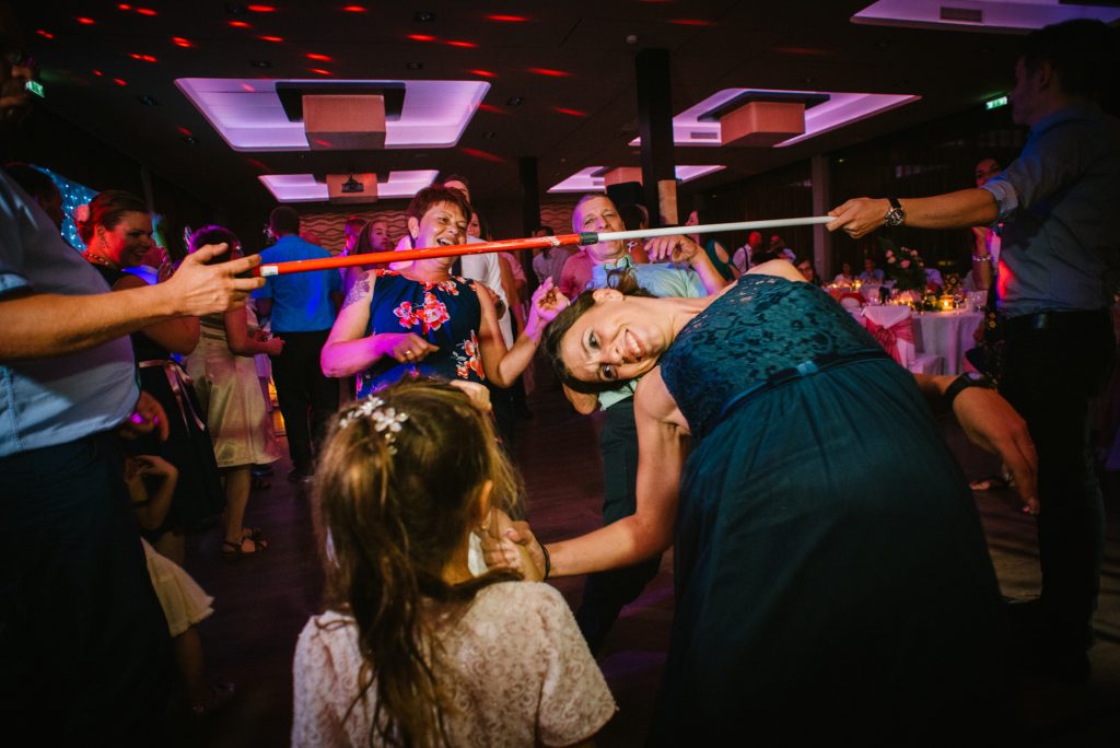 Wedding guest bending low under limbo stick while others cheer and laugh around.