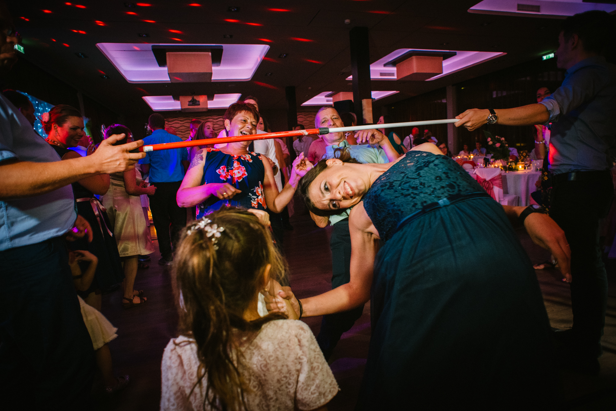Wedding guest bending low under limbo stick while others cheer and laugh around.