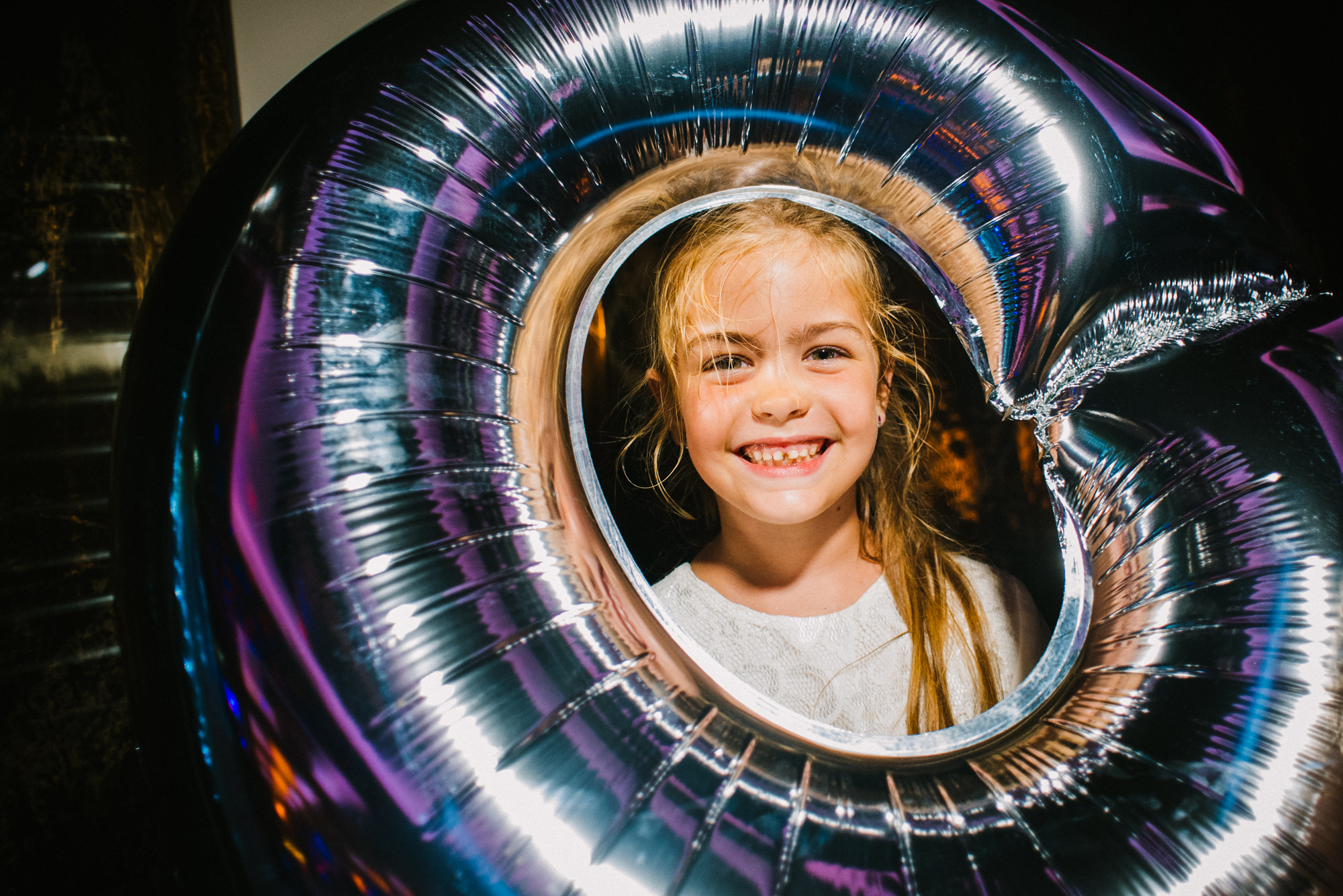 Young girl smiling through large silver balloon frame during wedding party celebration.