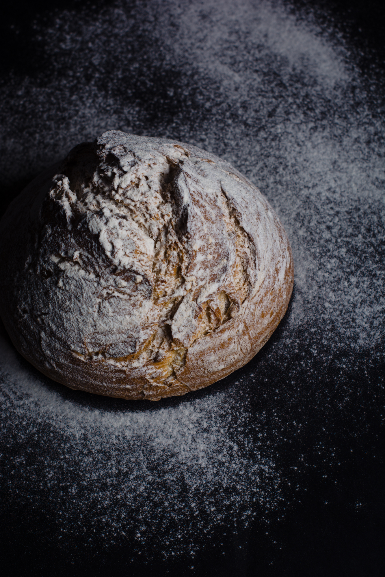 Rustic sourdough loaf dusted with flour on dark surface, moody bakery food photography.