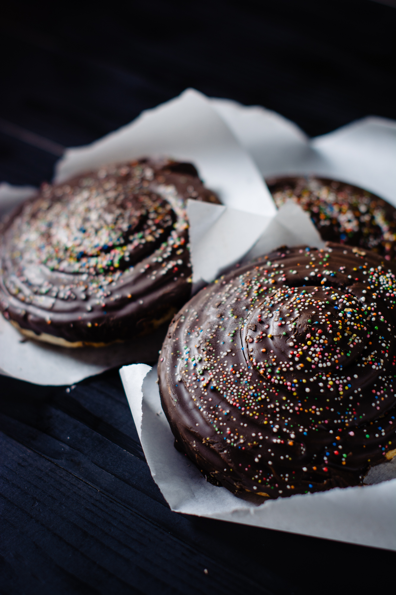 Chocolate pastries topped with colourful sprinkles on dark background, styled for bakery visuals.