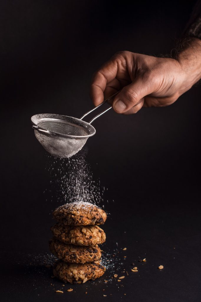Hand sifting powdered sugar over stacked oat cookies against dark background with dramatic lighting.