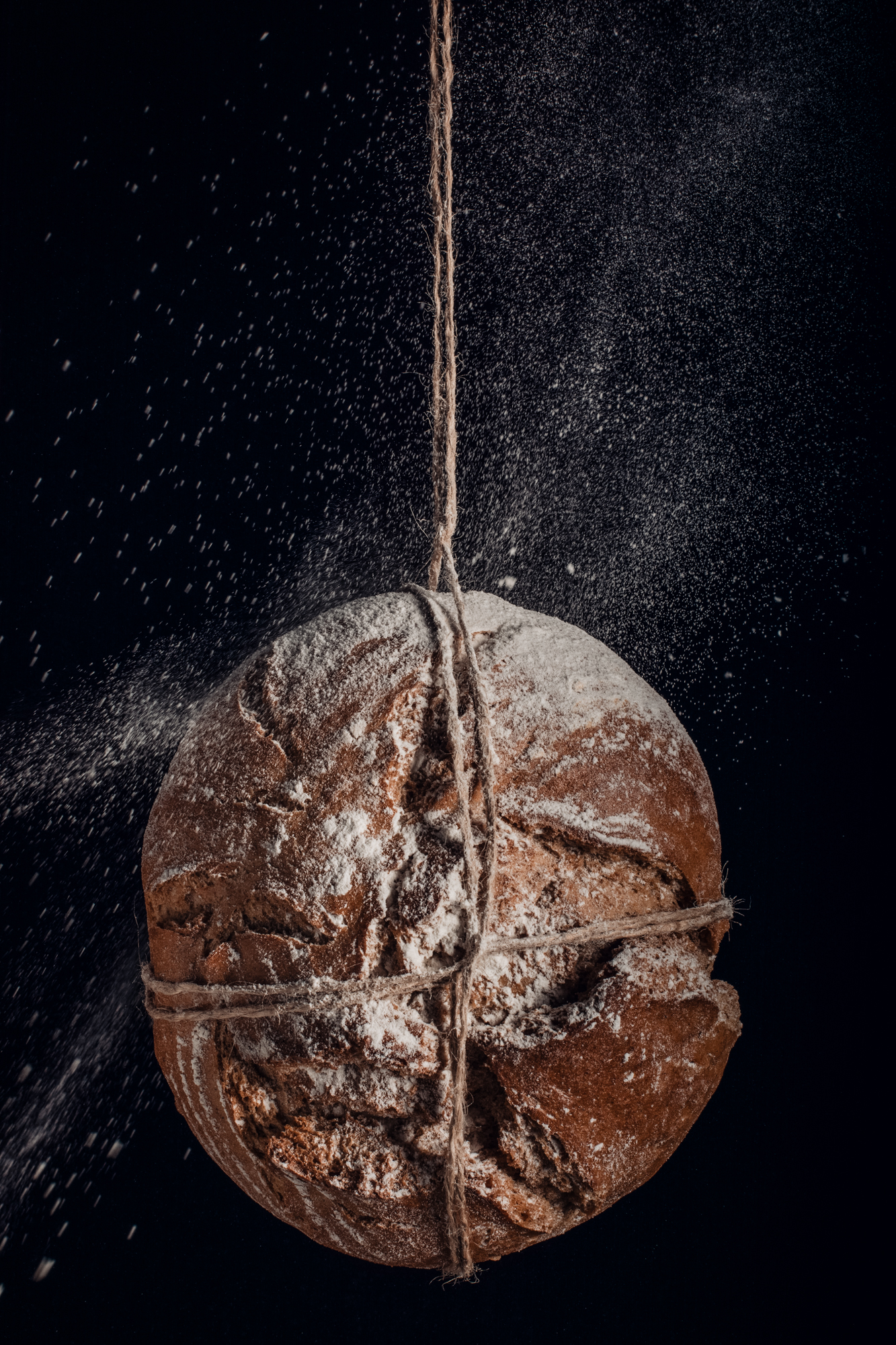 Flour falling over tied sourdough loaf, captured in motion with dramatic side lighting.