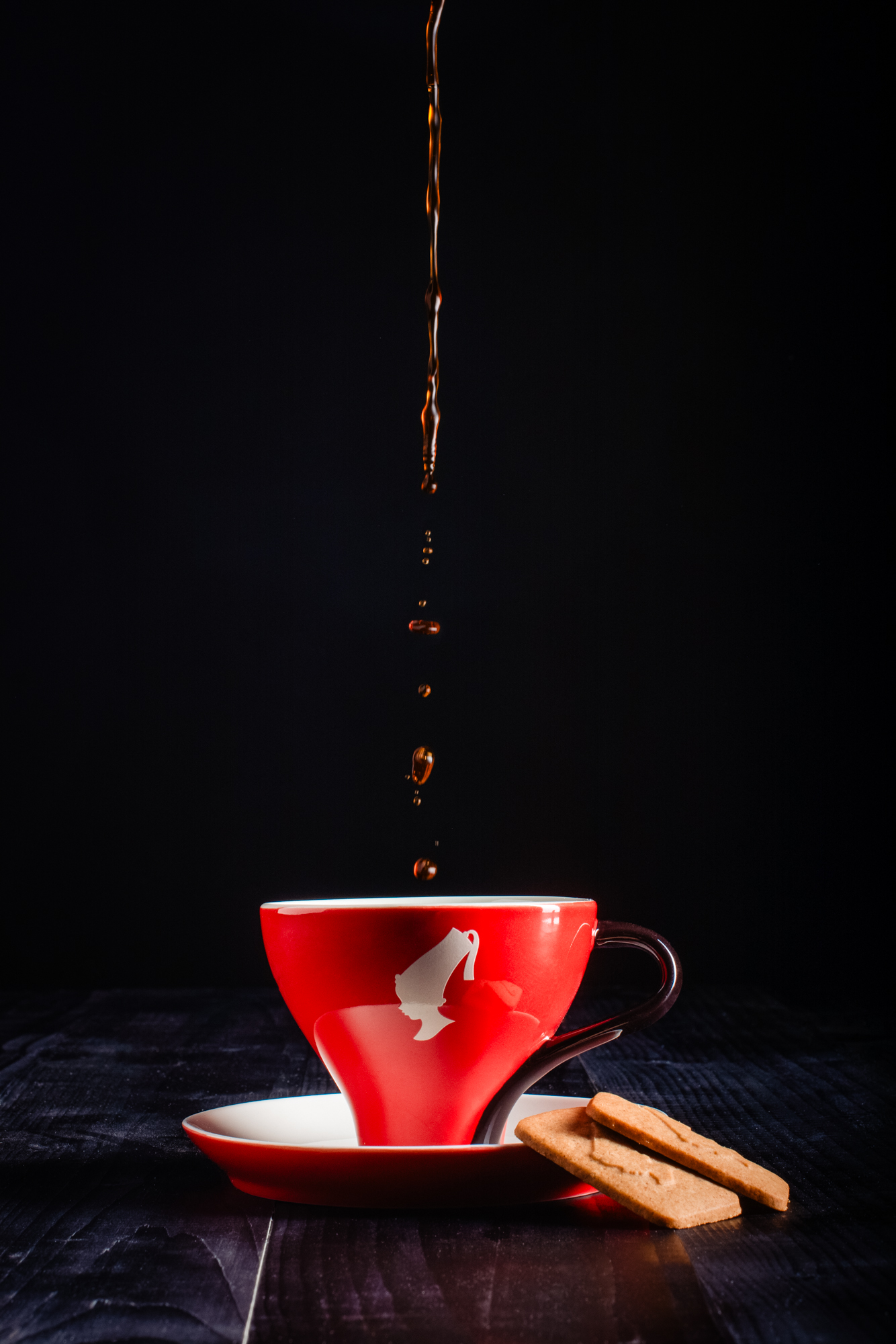 Coffee pouring into red cup with biscuit on saucer, styled for bakery branding photography.