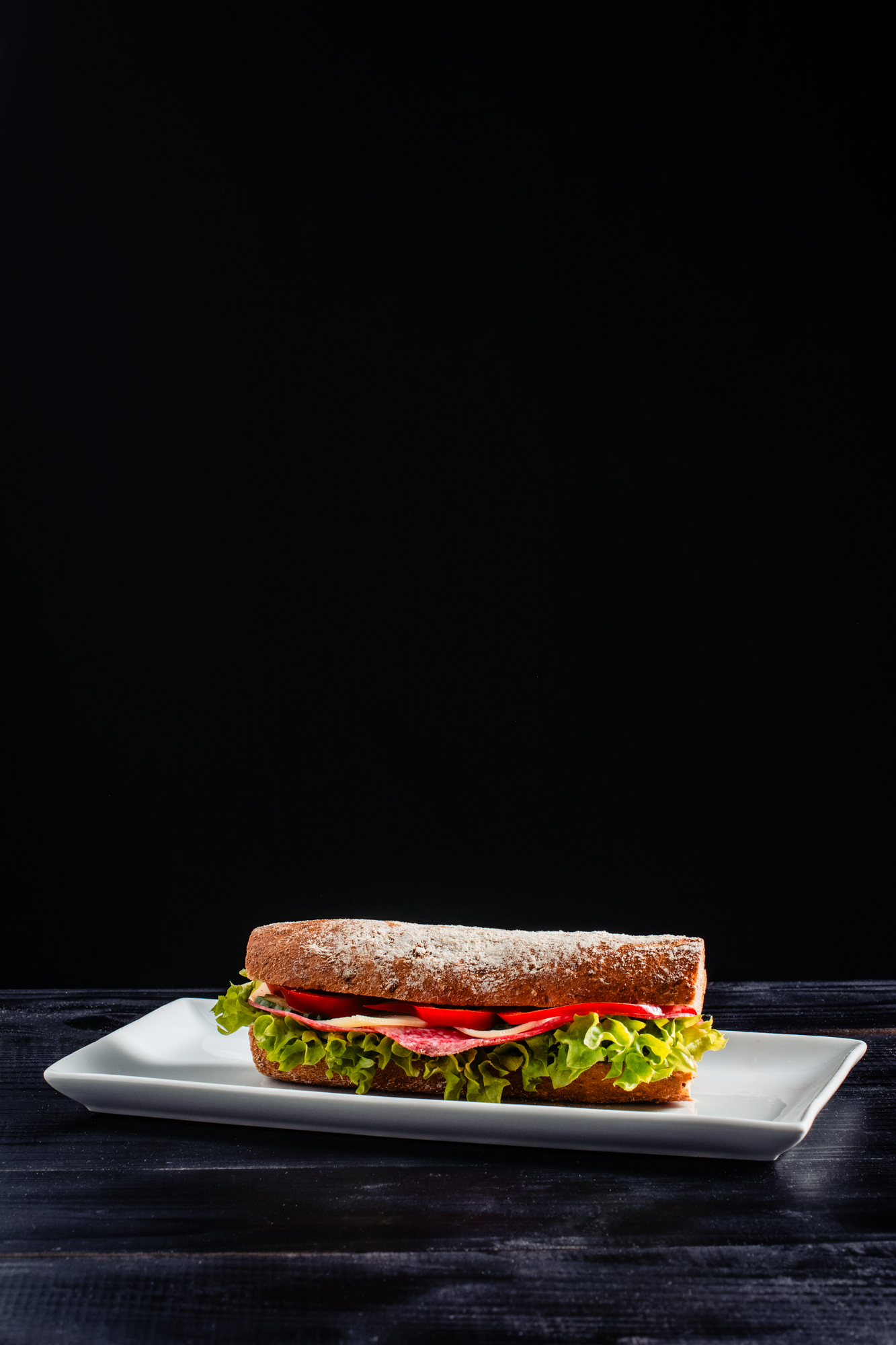 Fresh baguette sandwich with lettuce and salami on plate against clean dark background.