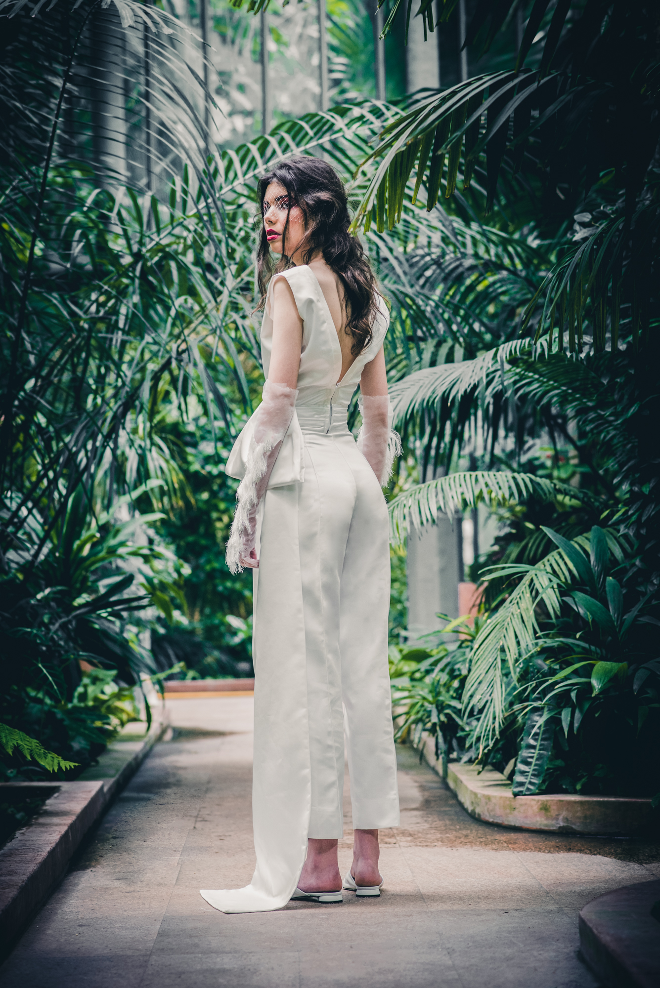 Bride in elegant fitted jumpsuit looking back in tropical greenhouse Budapest.