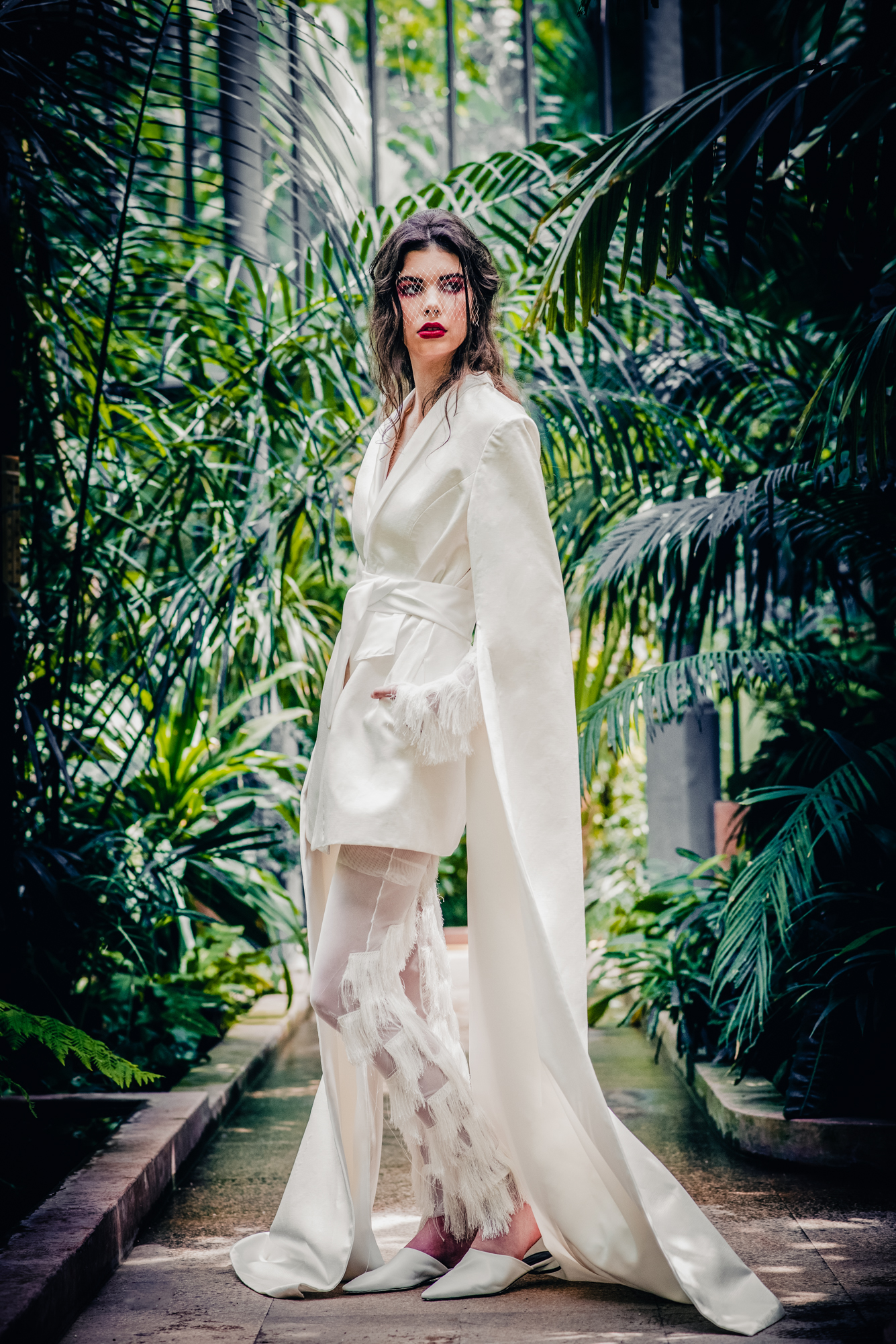Bride in modern white outfit posing among lush greenhouse plants in Budapest botanical garden.