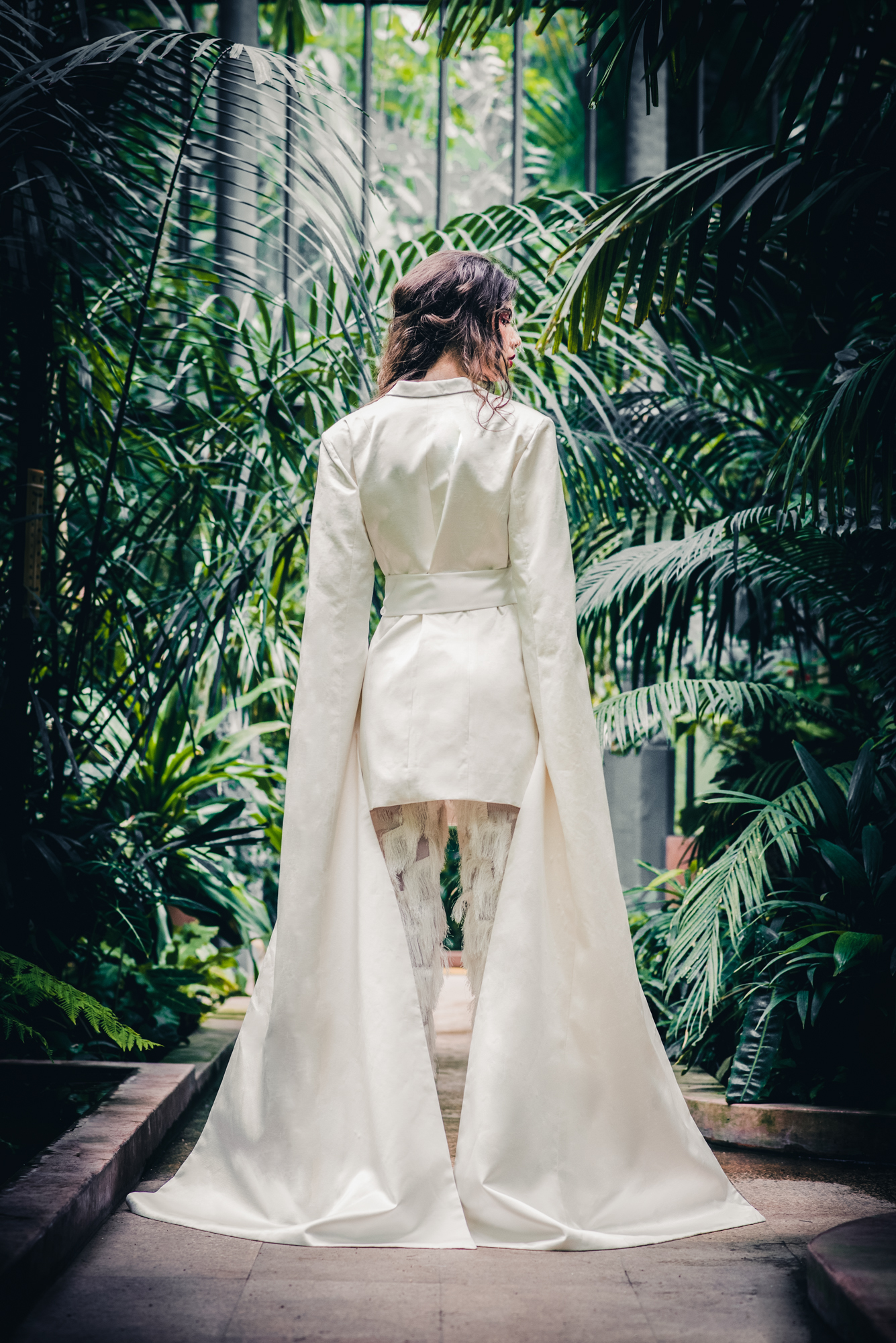 Back view of bride in long white tailored coat walking through ELTE Füvészkert greenhouse.