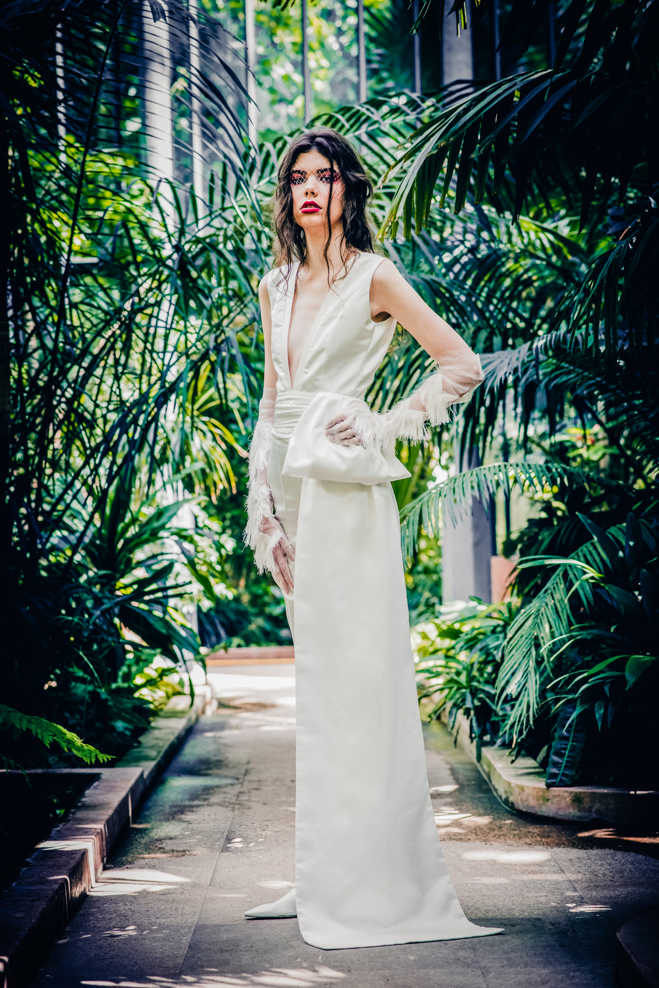 Fashion bridal pose in sleeveless white dress with feather details in botanical greenhouse.