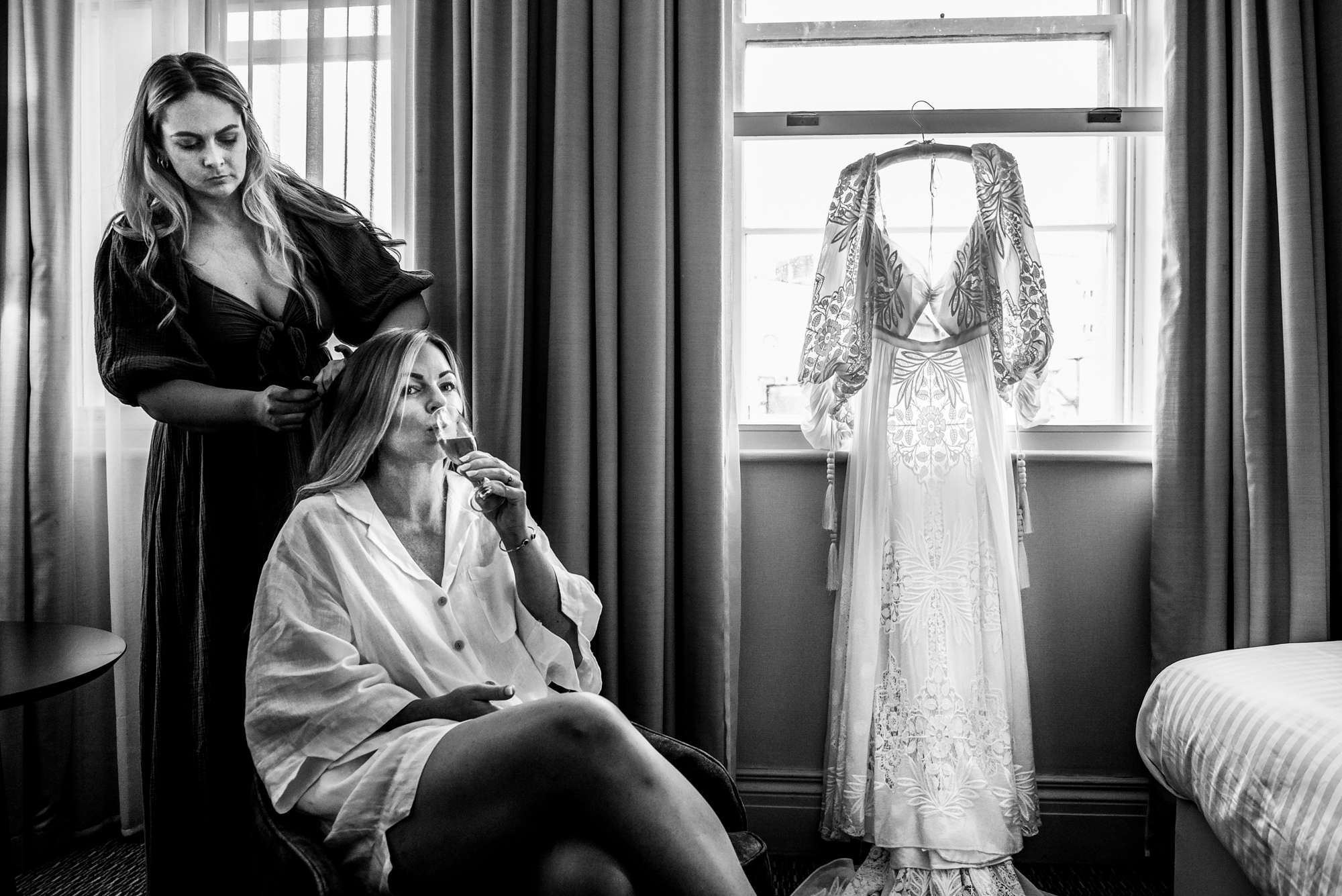 Bride sipping champagne while getting hair done beside wedding dress in window at Bristol Marriott Royal Hotel.