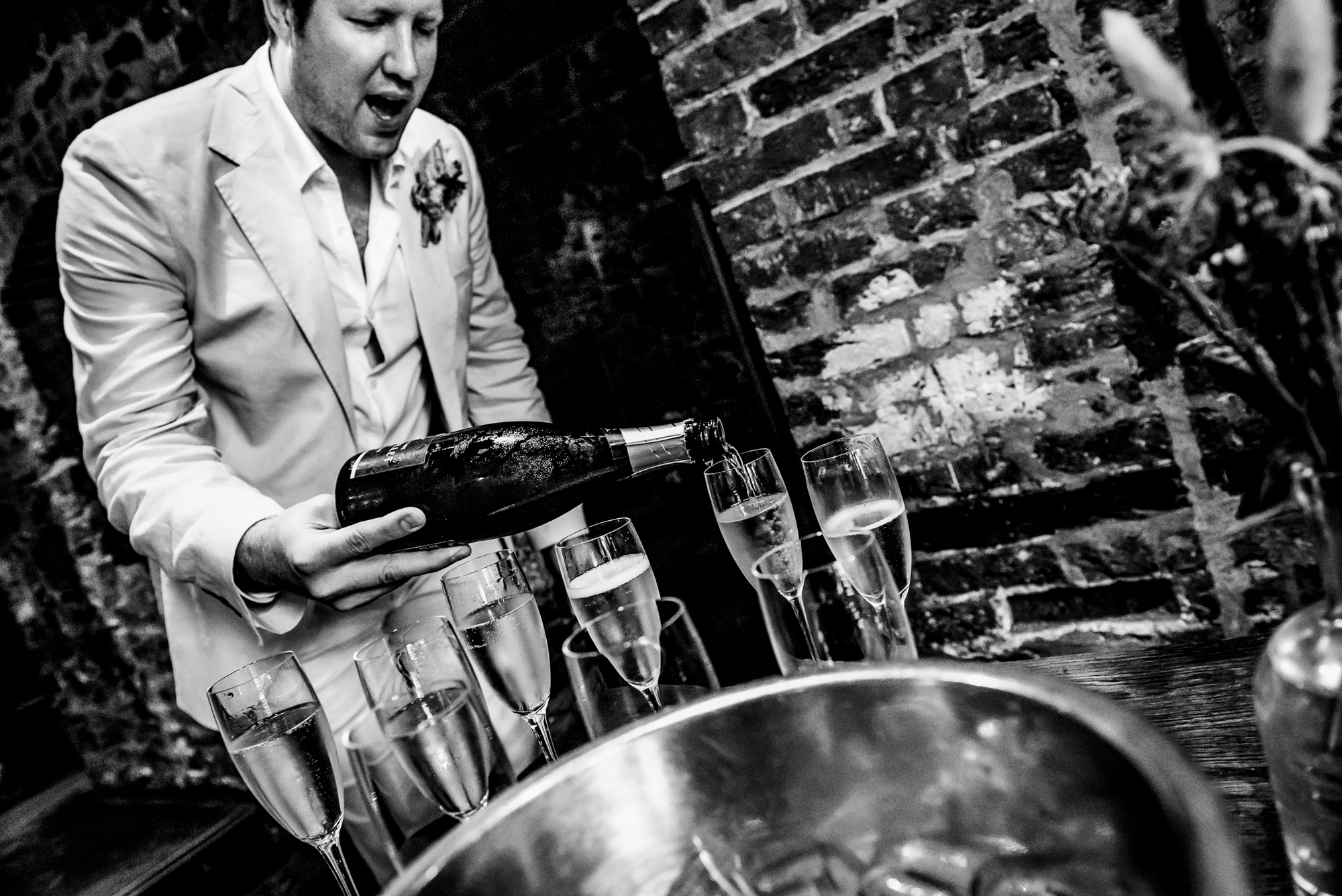 Wedding guest opening champagne surrounded by glasses in atmospheric brick cellar setting.