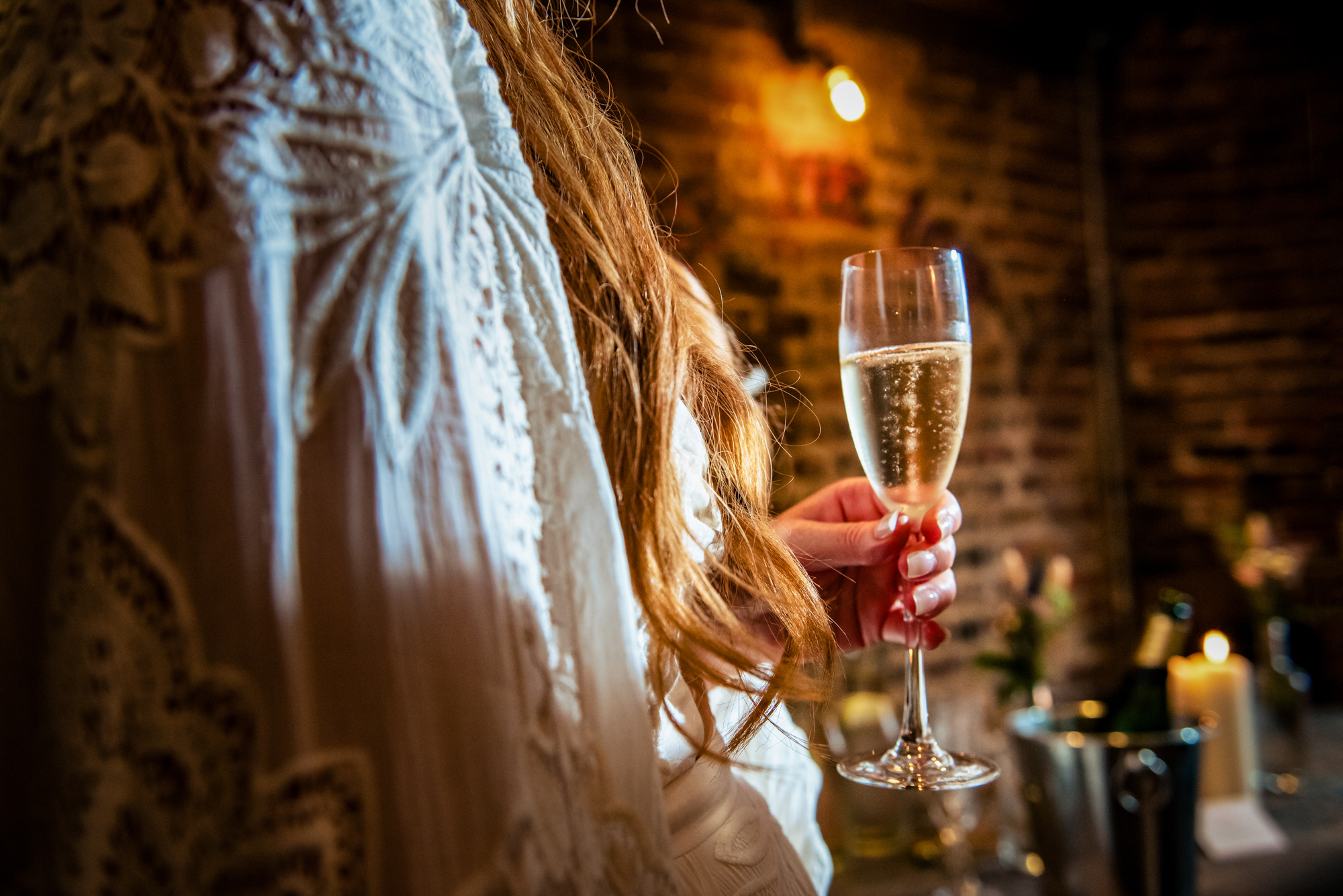 Bride holding champagne glass with soft light and textured brick background.