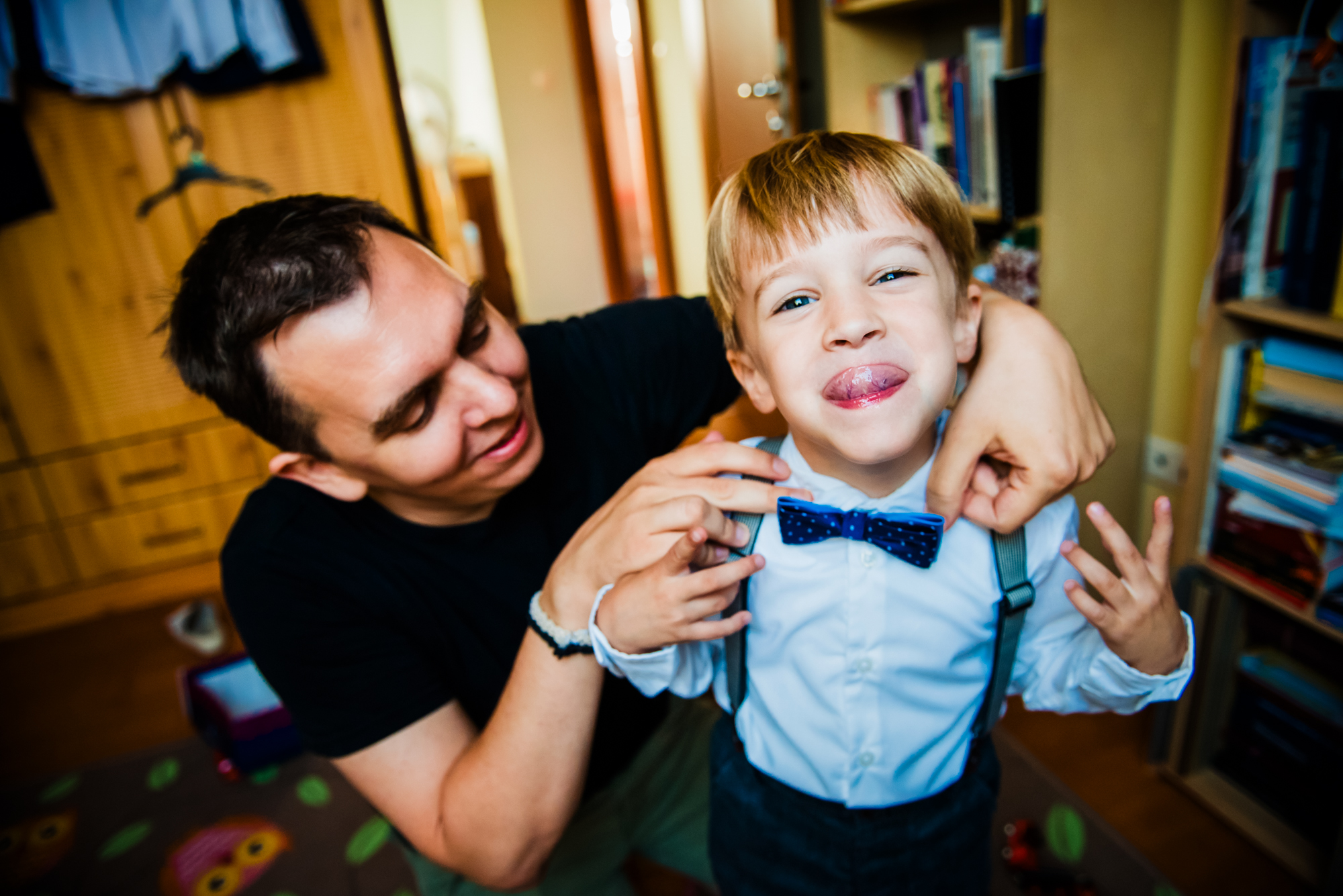 Father adjusting young boy’s blue bow tie during christening morning preparations at home.