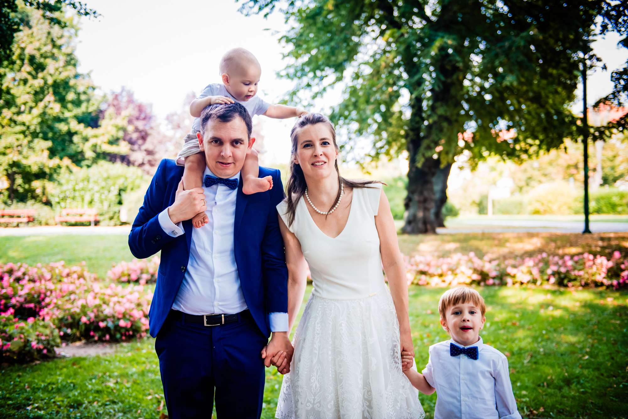 Family walking through a flower-filled park after a christening, with toddler riding on father’s shoulders.