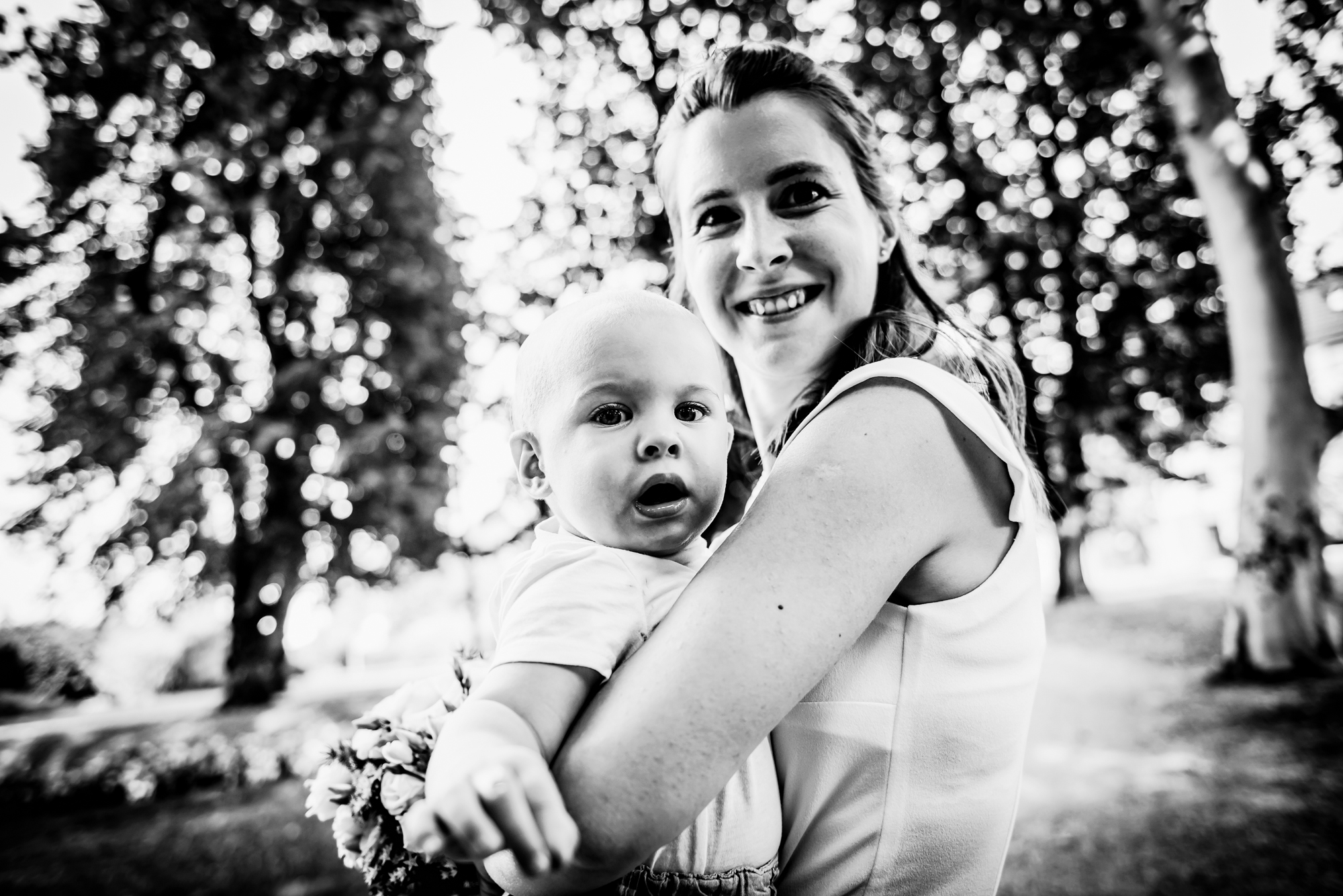 Mother holding her baby during an outdoor christening portrait session beneath tall trees.