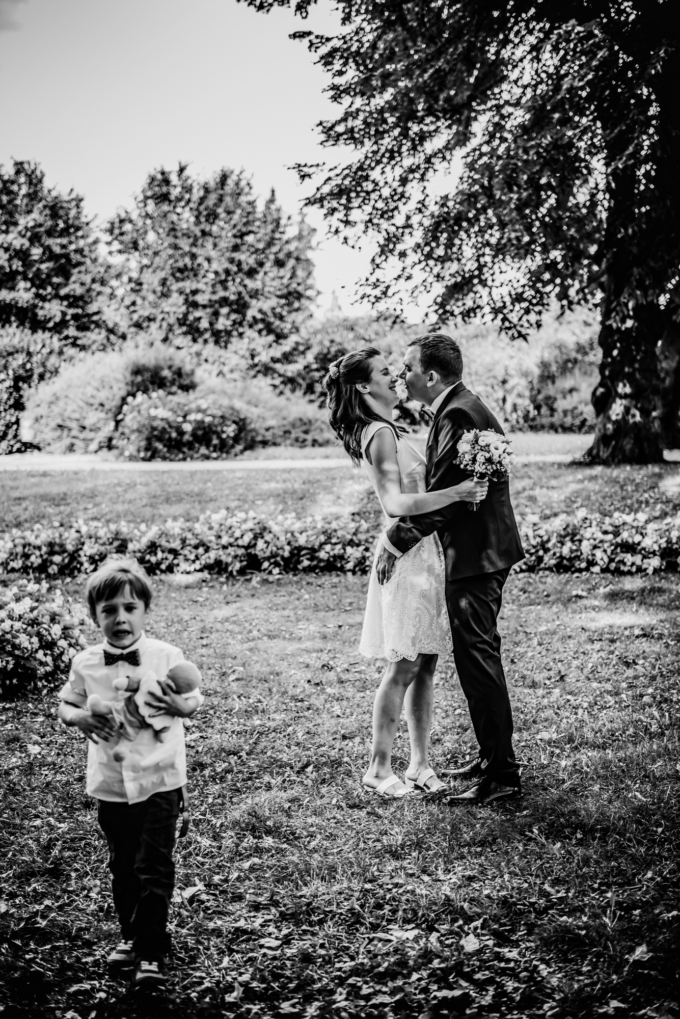 Young boy holding a teddy bear while parents kiss during relaxed outdoor family christening photos.