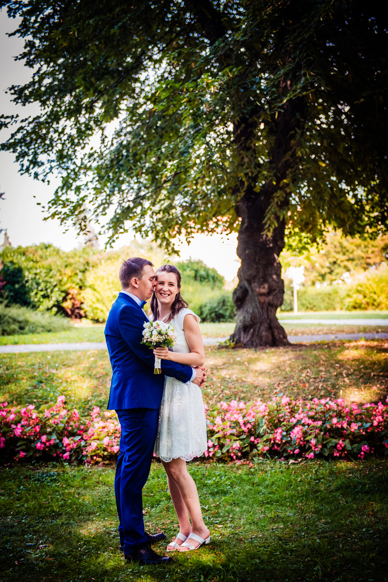 Couple embracing beneath a large tree during a sunny park christening portrait session with pink flowers behind them.