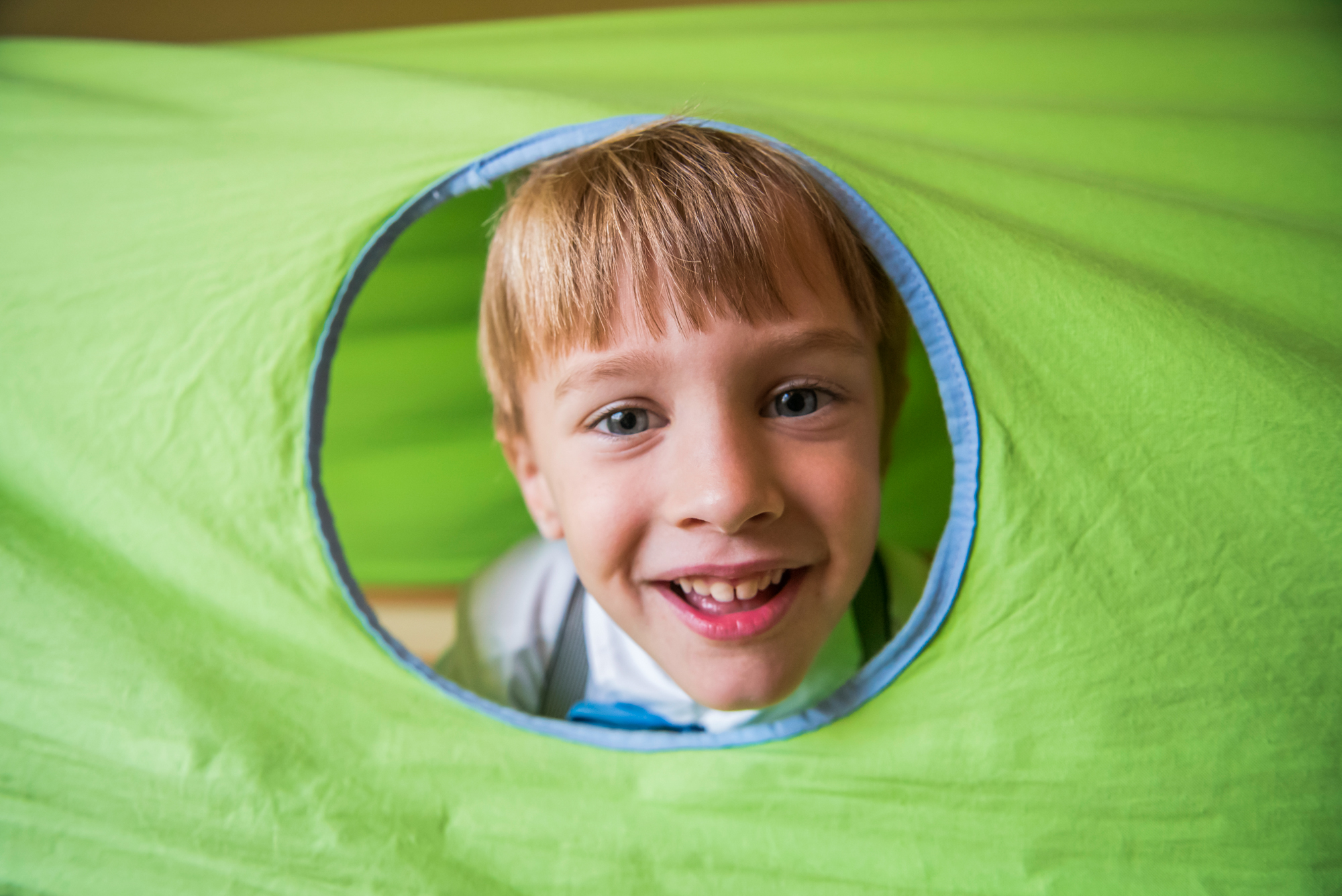 Laughing little boy peeking through a bright green play tunnel while dressed for his christening.