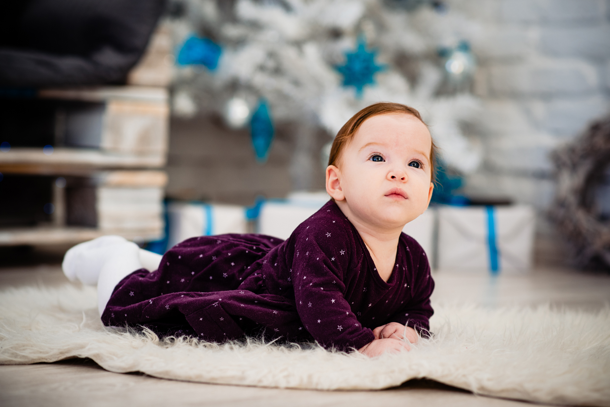Baby Bella lies on a soft rug in front of a white Christmas tree with blue decorations.