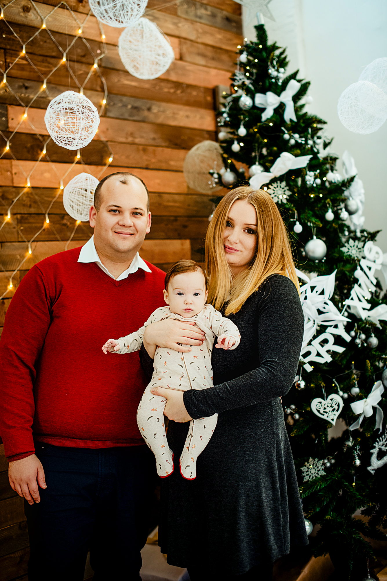 Family portrait with baby Bella beside a decorated Christmas tree and warm festive lights.