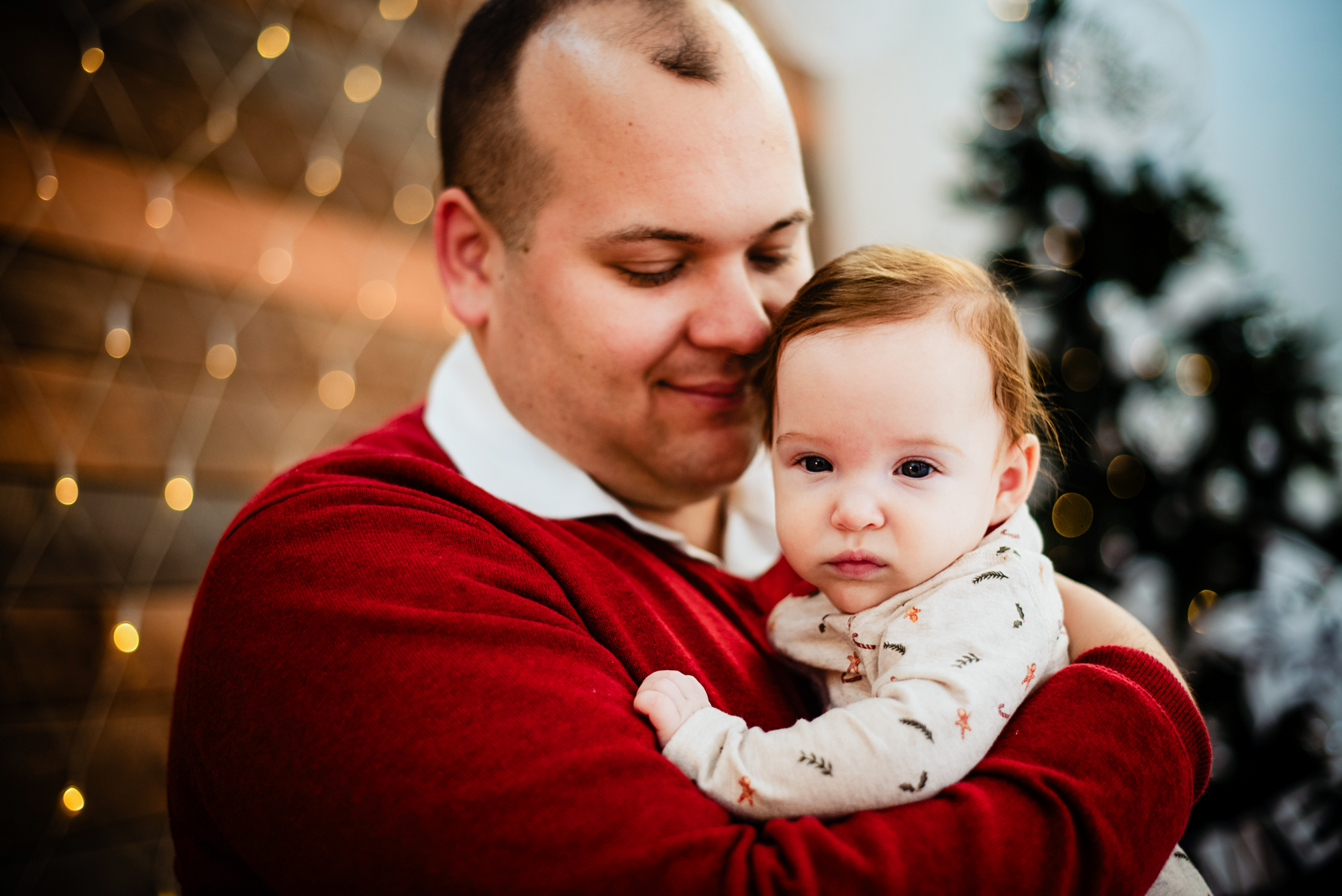 Father cuddles baby Bella in front of soft Christmas lights during festive family session.