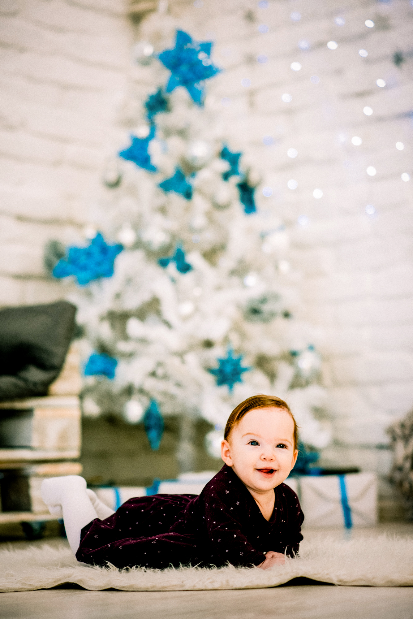 Baby Bella smiles on a fluffy rug during a Christmas studio session with festive tree behind.