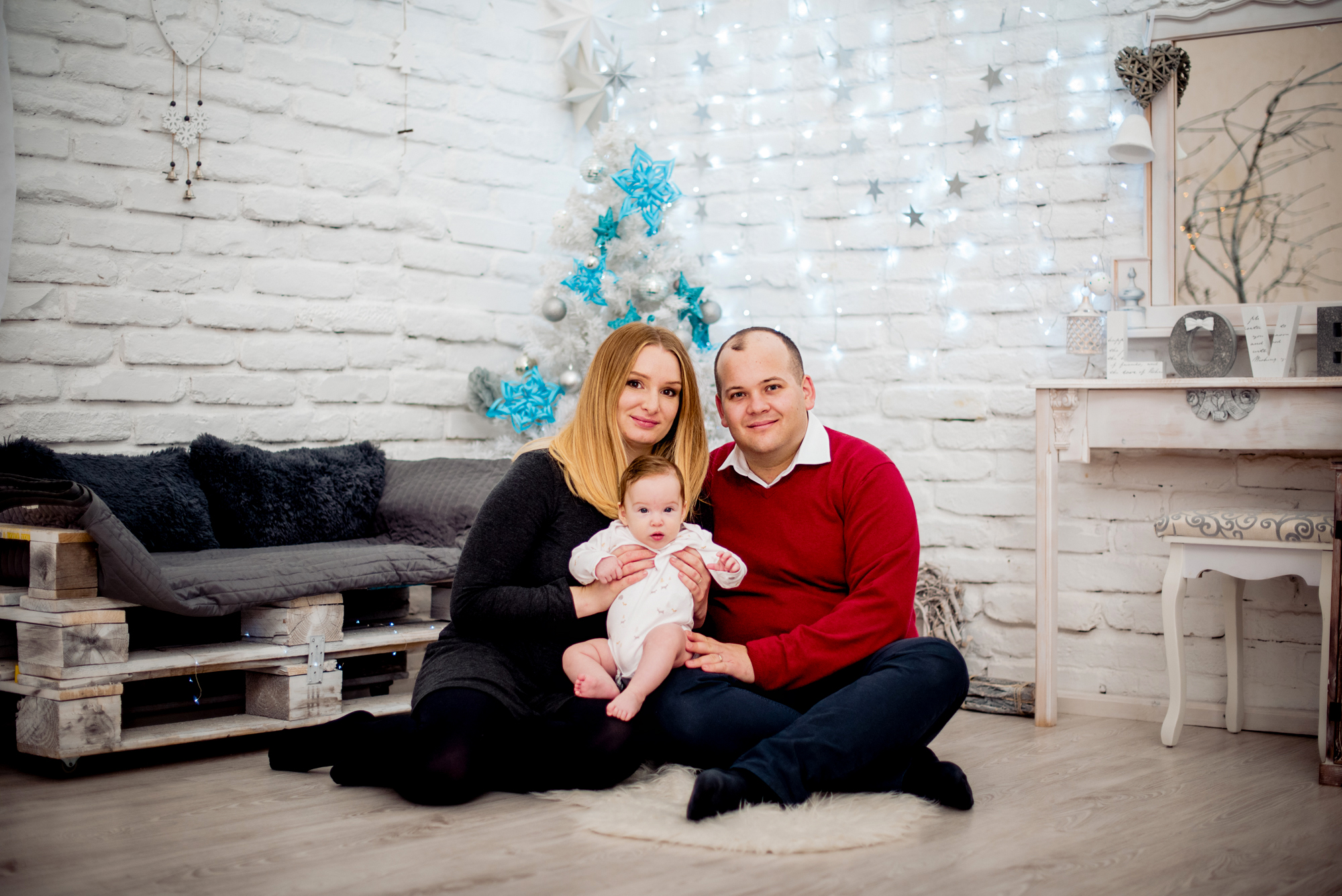 Family portrait beside a white Christmas tree with blue decorations and soft fairy lights.