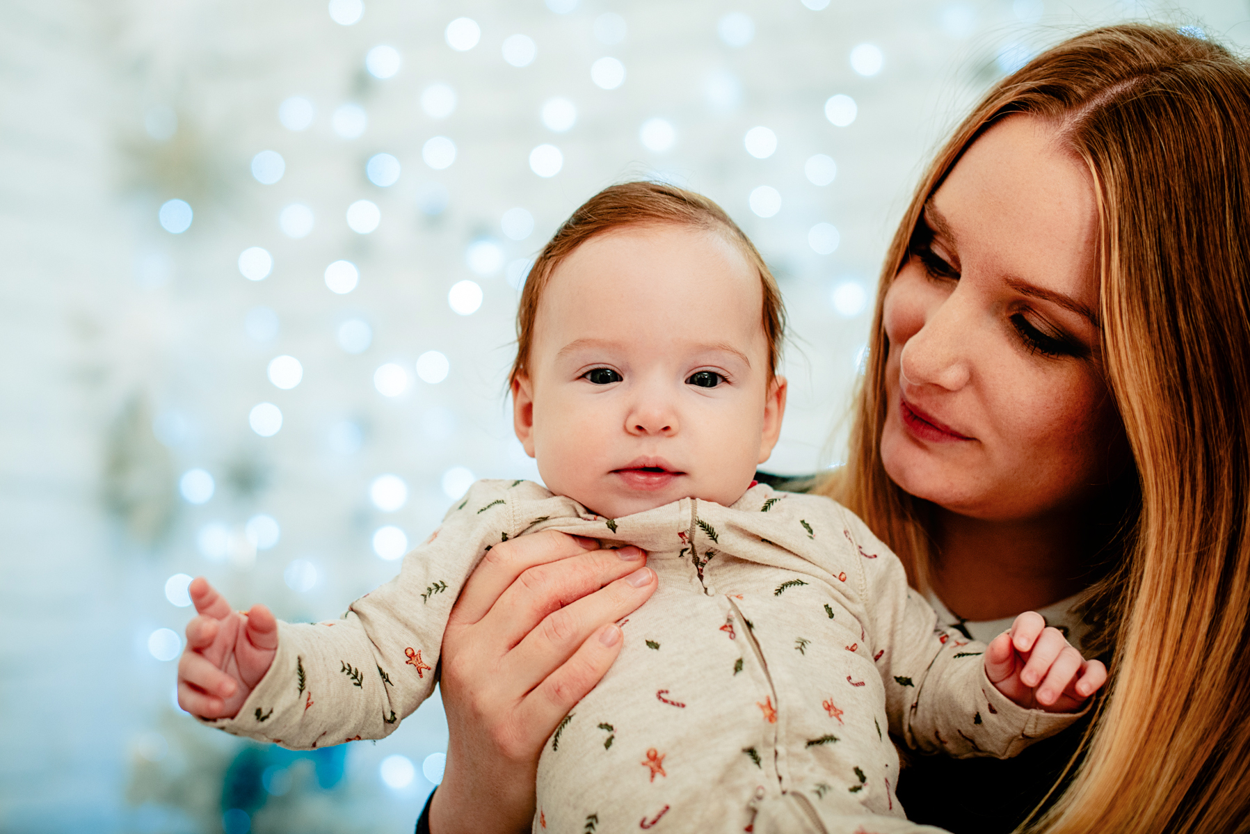 Mother holds baby Bella close in front of soft Christmas lights and festive decor.