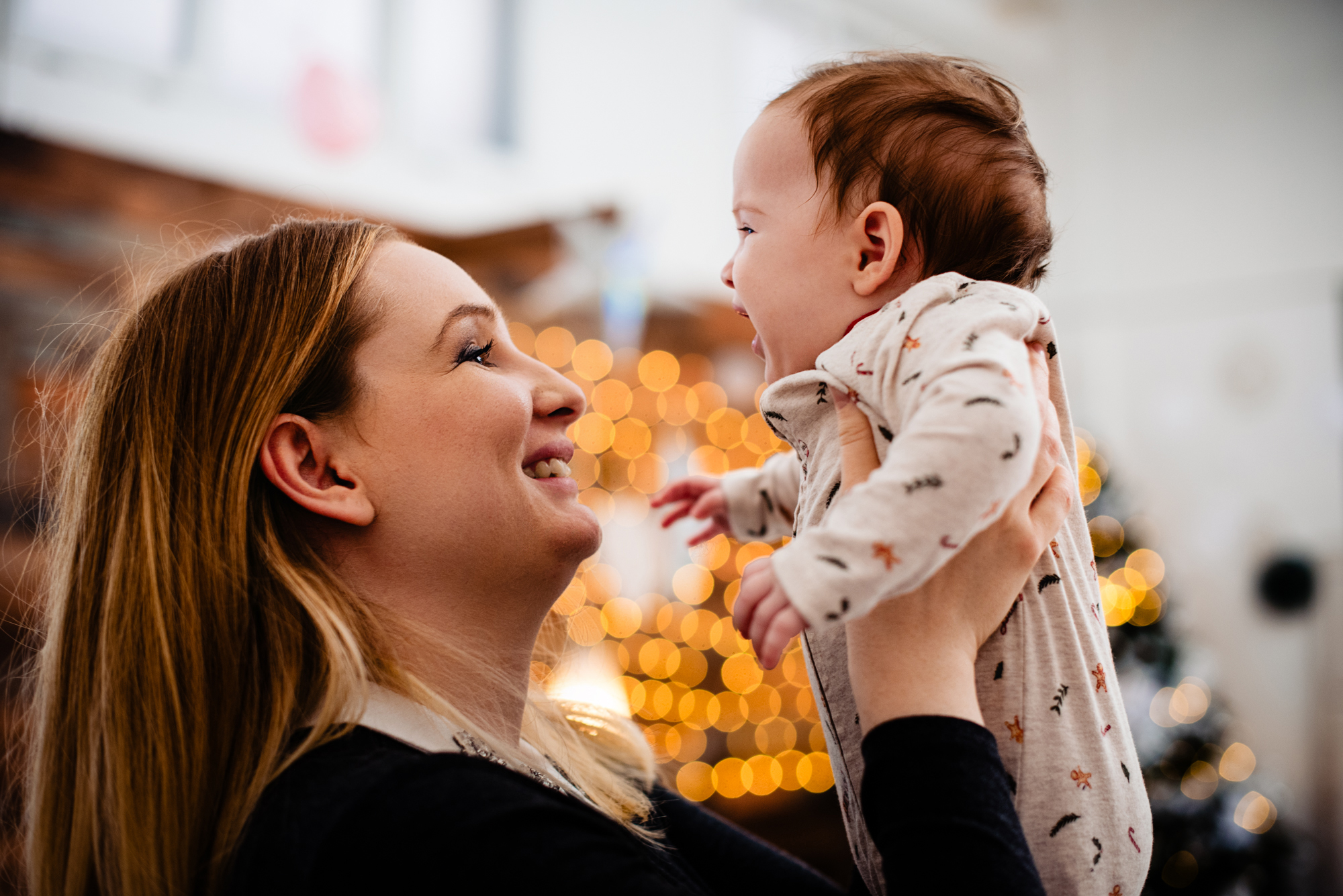 Mother lifts baby Bella while they smile at each other in front of warm Christmas lights.