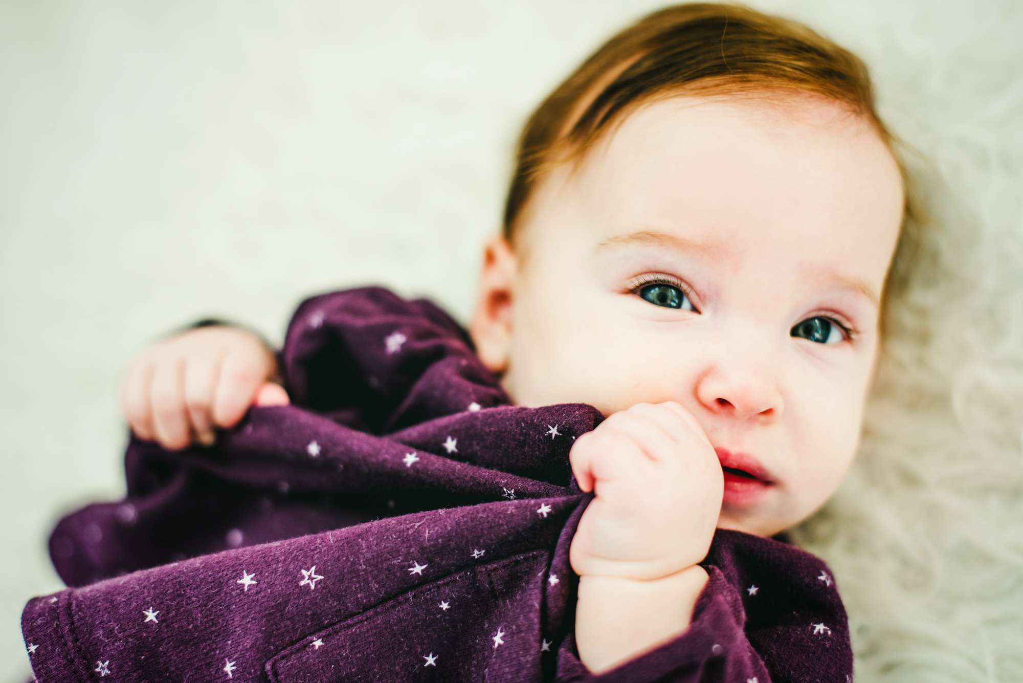 Close-up of baby Bella chewing her dress while lying on a soft white rug.