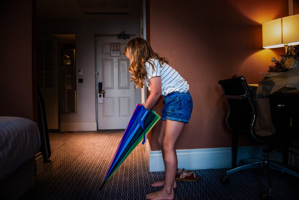 Little girl picking up colourful umbrella in softly lit hotel room before ceremony at Bristol Marriott Royal Hotel in Bristol.