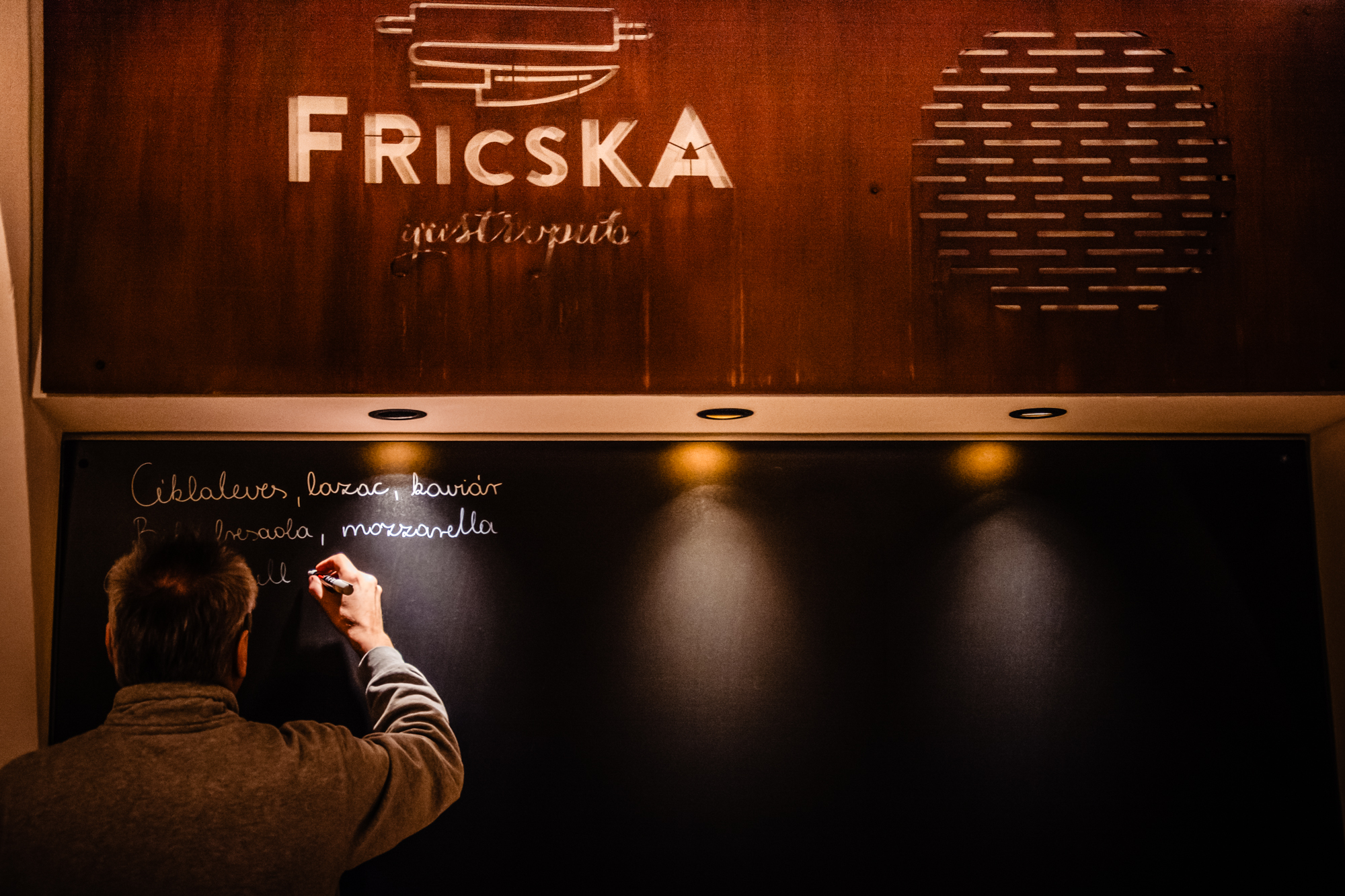 Chef writing daily specials on chalkboard beneath Fricska Gastropub sign in Budapest restaurant interior.