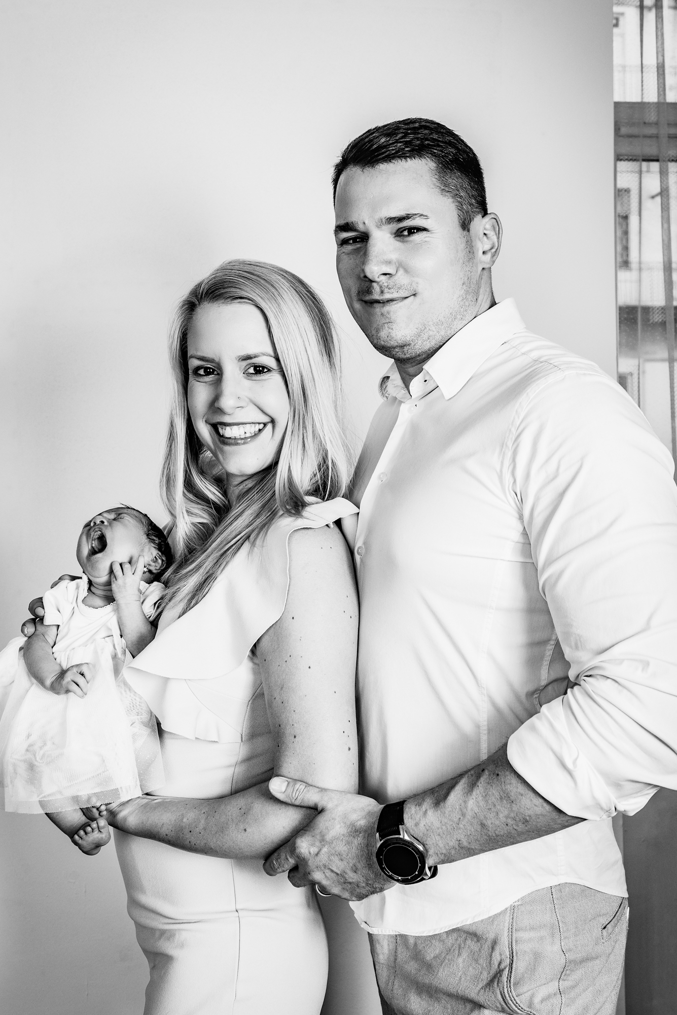 Mother smiling while holding newborn baby Kendra, father standing close during home newborn session.