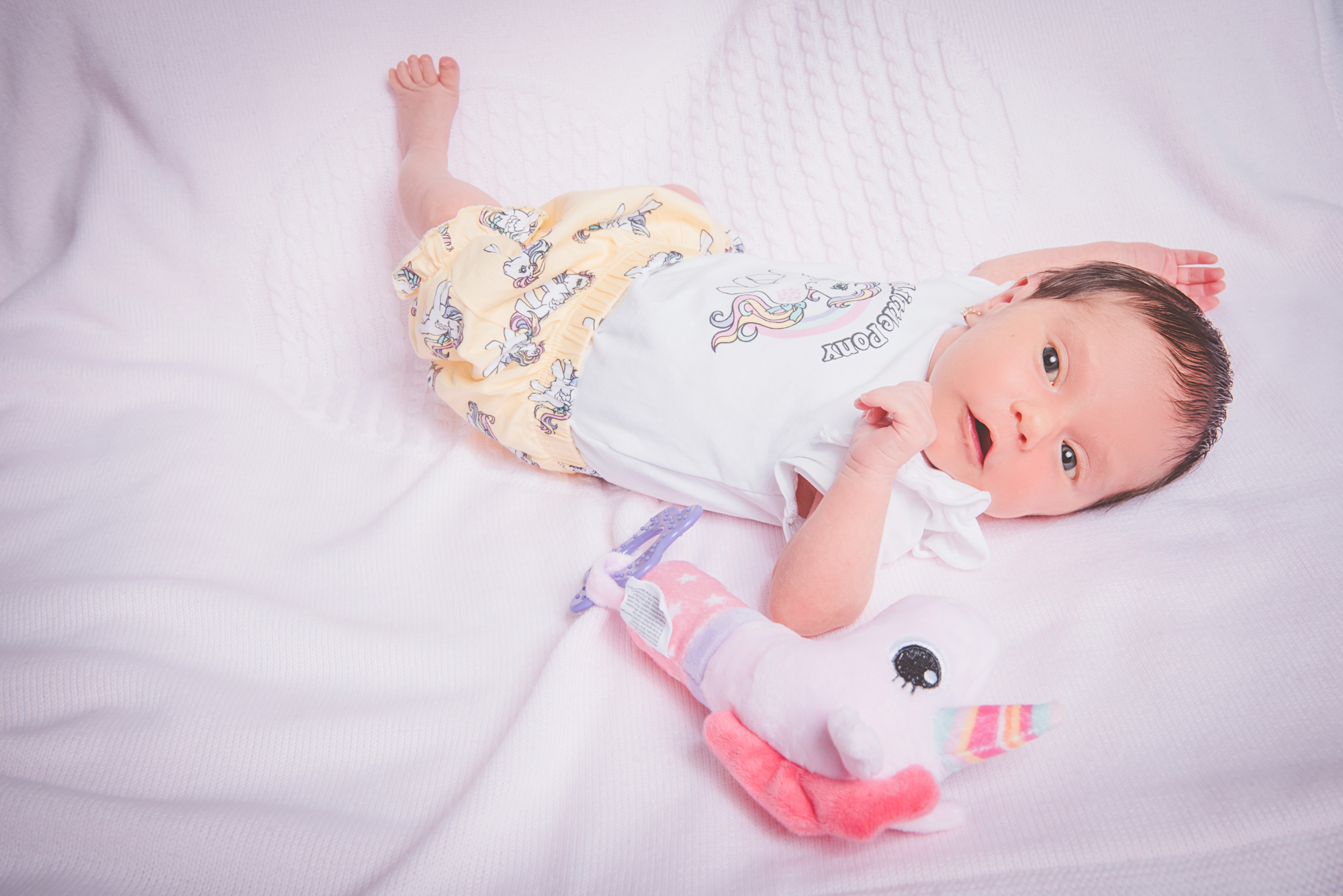Newborn baby Kendra lying awake on soft blanket with toy during relaxed home session.