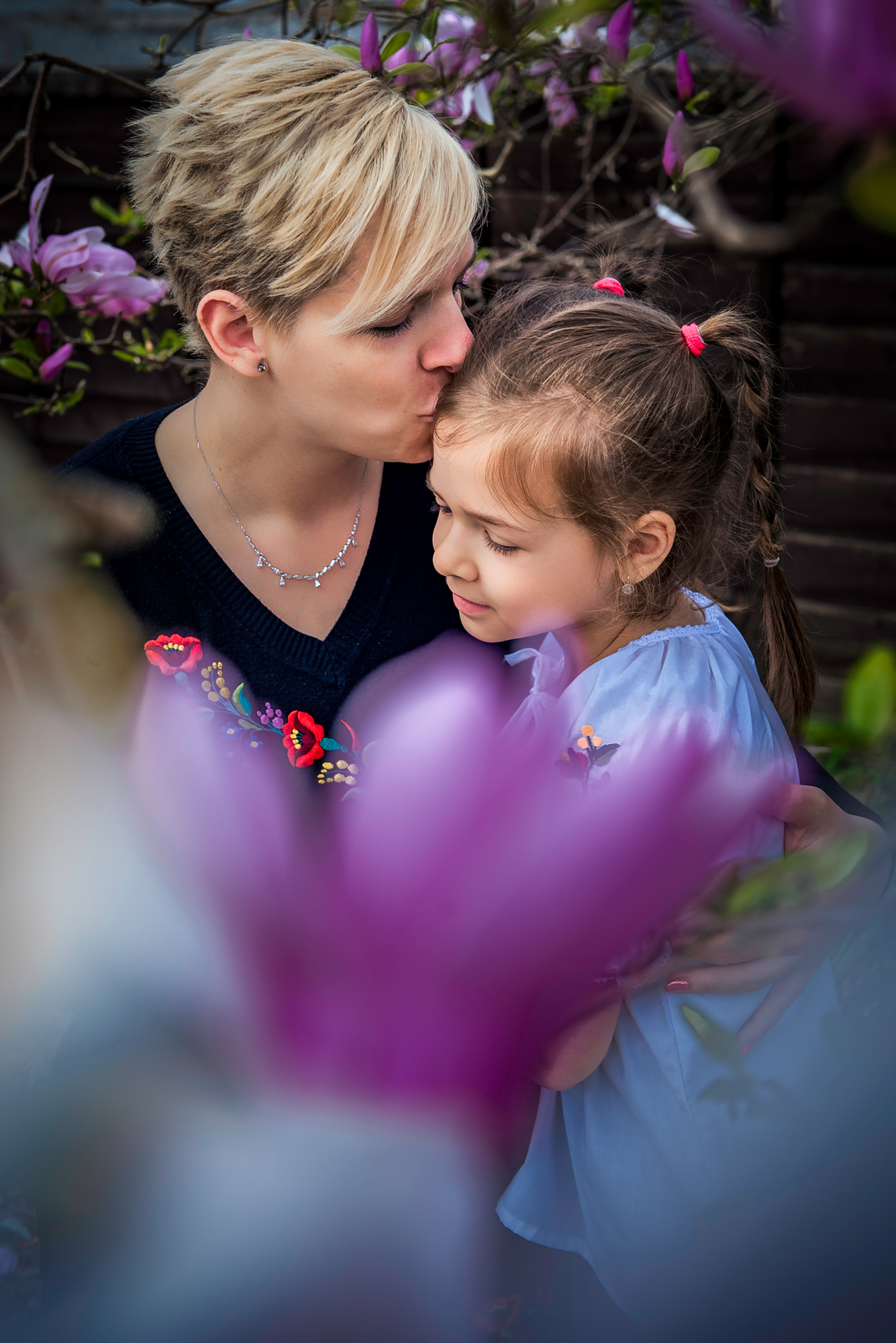 Mum kisses her daughter’s head among pink blossom in a spring family portrait.