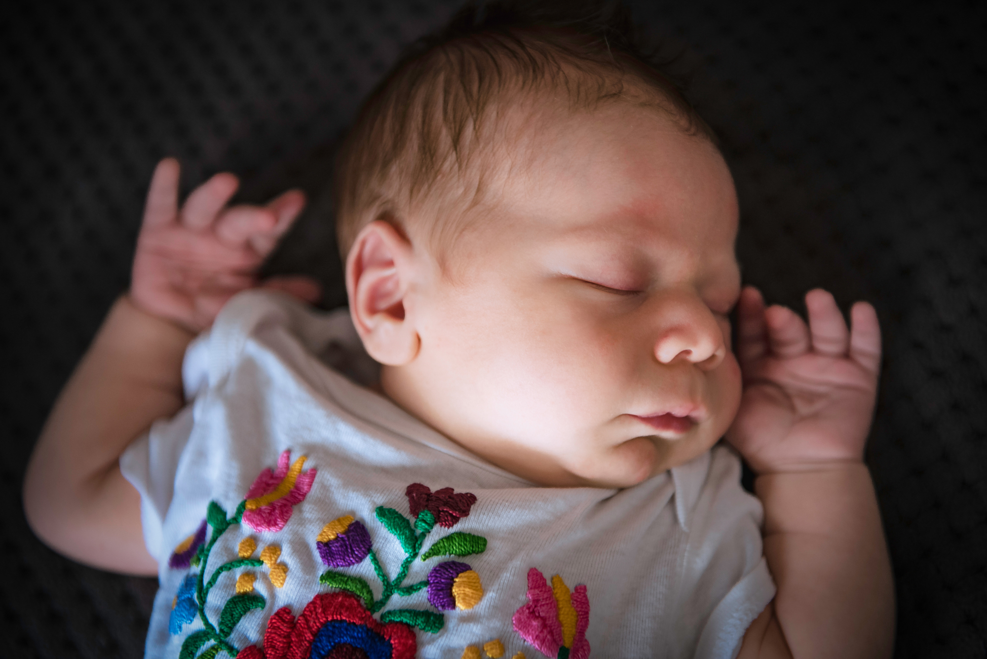 Sleeping newborn in an embroidered outfit lies on a dark textured blanket.