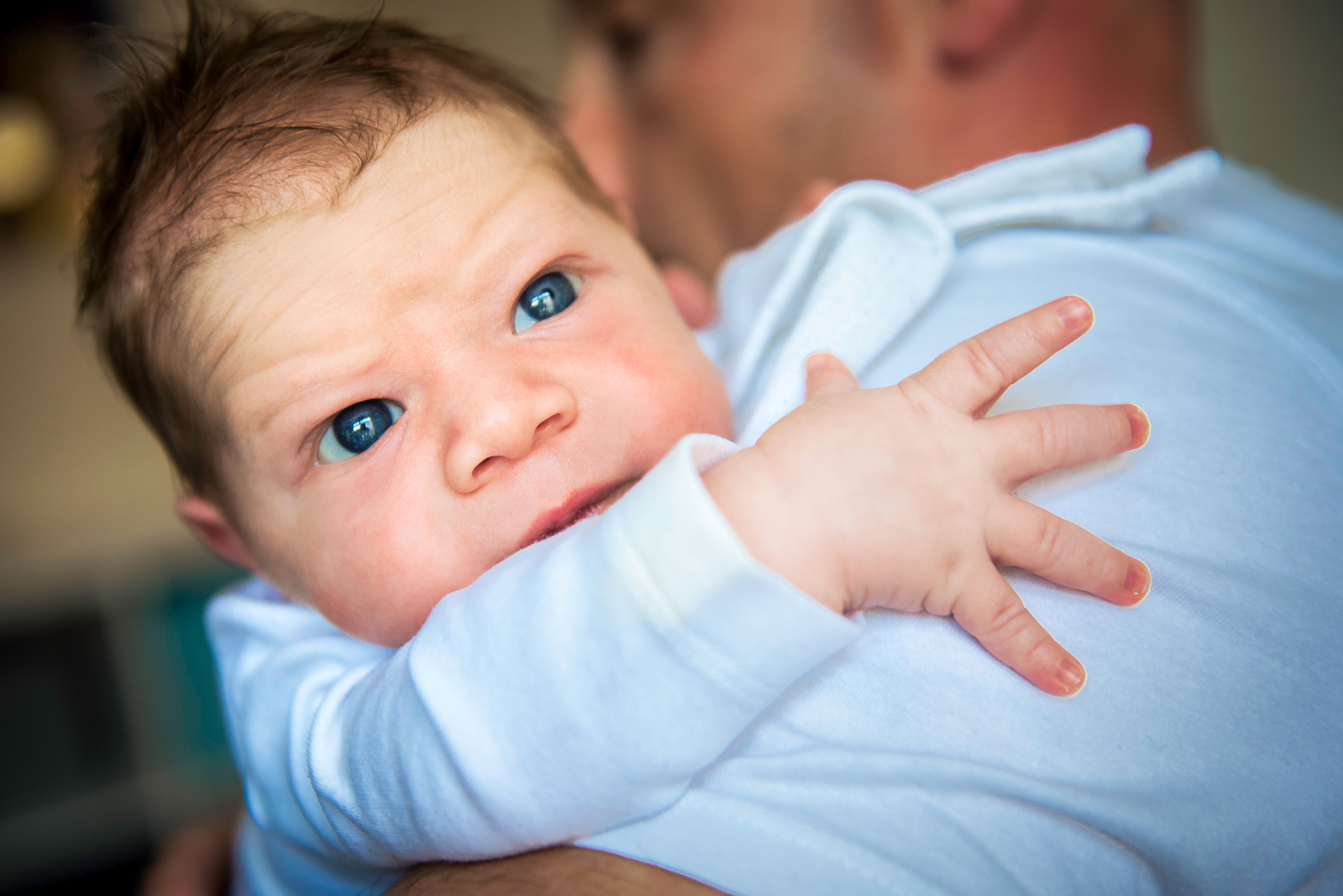 Wide-eyed newborn looks toward the camera while being held against dad’s shoulder.