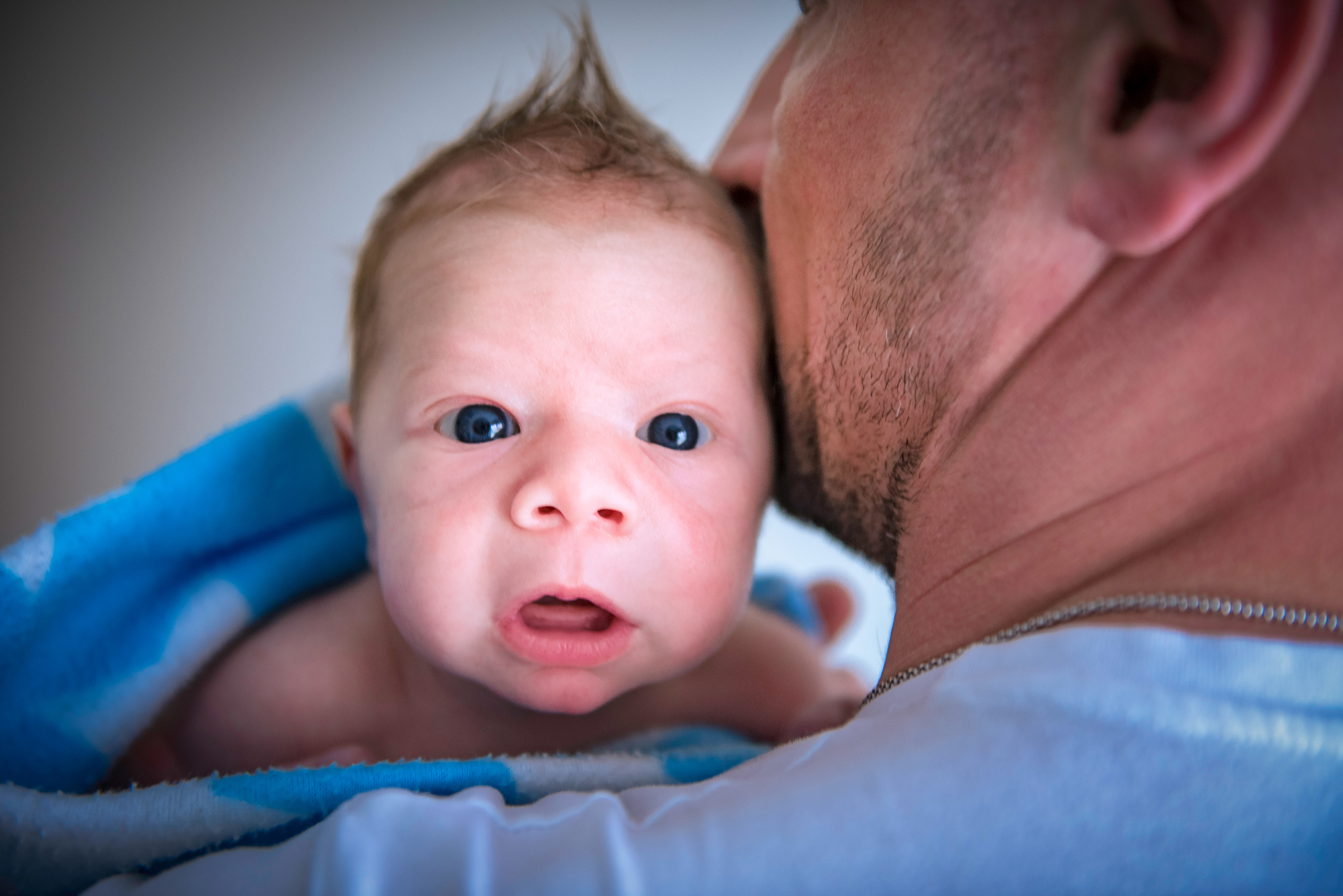 Blue-eyed newborn wrapped in a towel rests against dad after bath time.