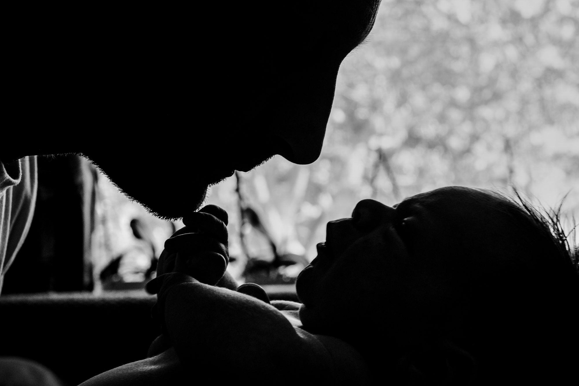 Silhouette of dad and newborn facing each other by a bright window.