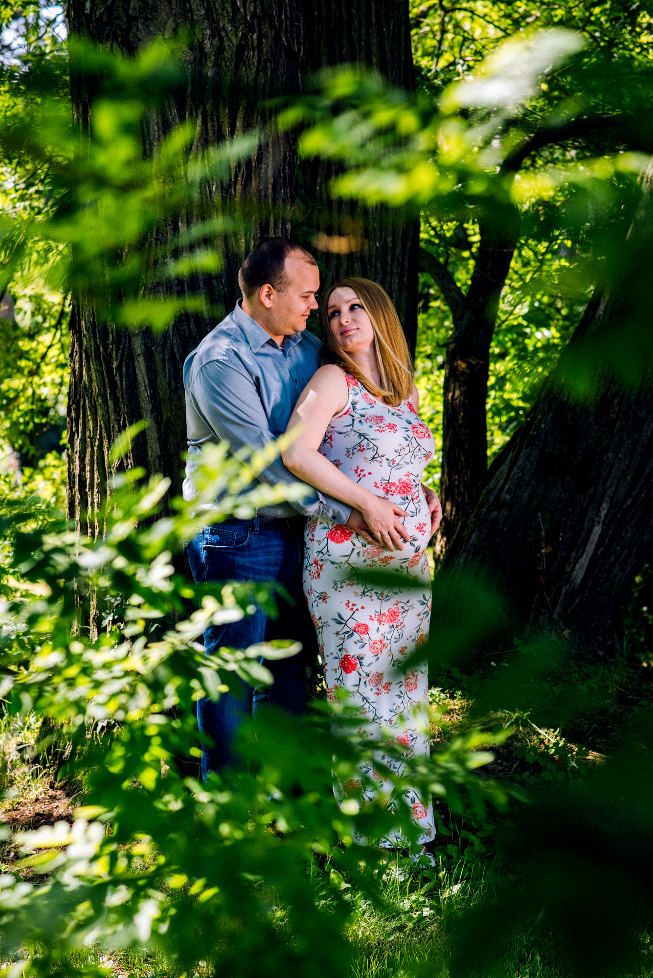 Couple embracing beneath trees at Vácrátót Botanical Garden, surrounded by soft green foliage at a maternity photoshoot.